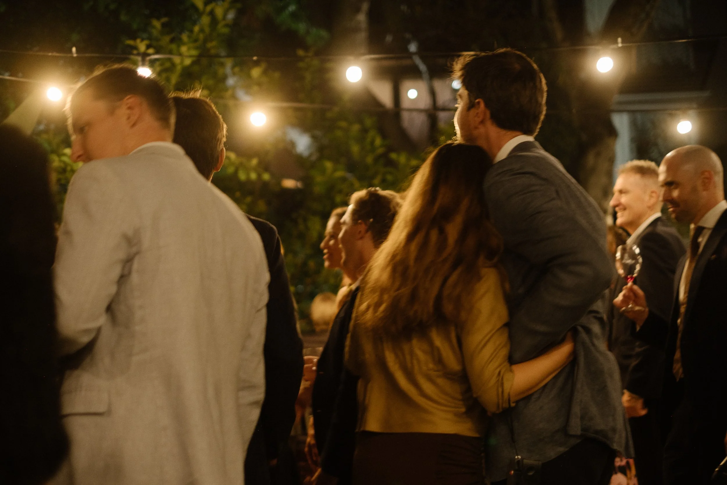 People at an outdoor evening event, some embracing, with string lights hanging above.