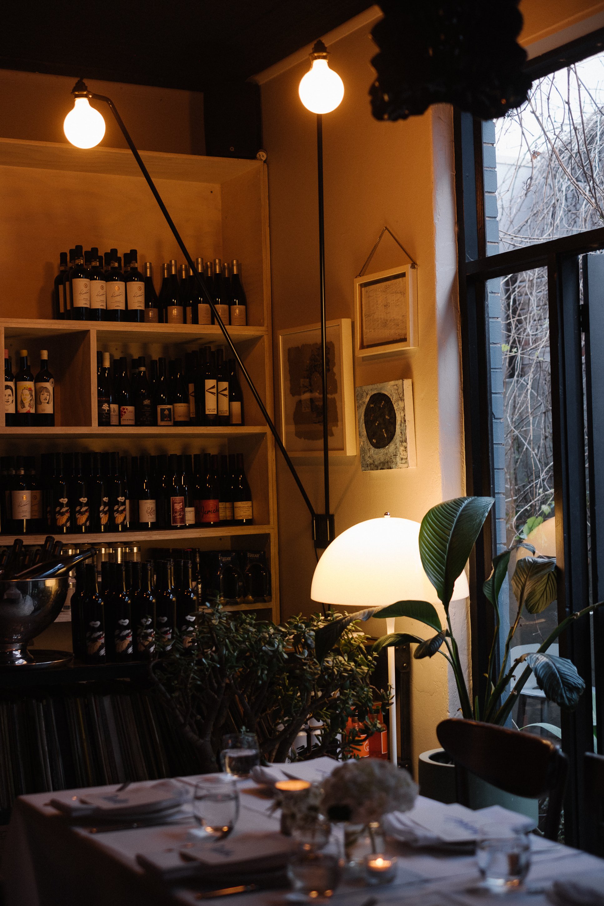 A cozy restaurant interior with shelves filled with wine bottles, a large window showing a winter scene outside, and warm lighting from hanging bulbs and a table lamp, with a dining table set with plates, glasses, and a floral centerpiece.