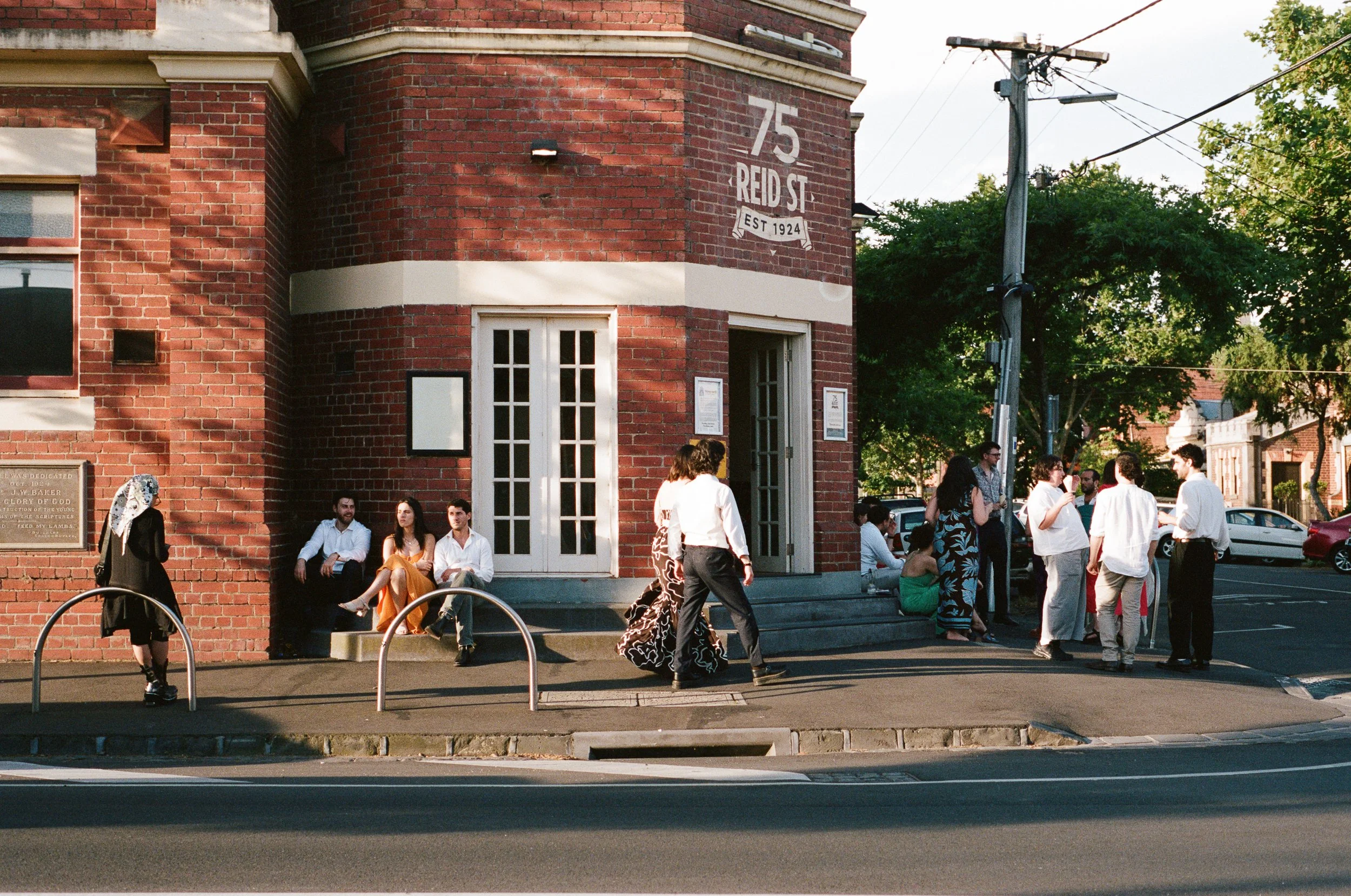 Group of people sitting and standing outside a red brick building on a street corner, with trees and parked cars in the background, during the late afternoon.