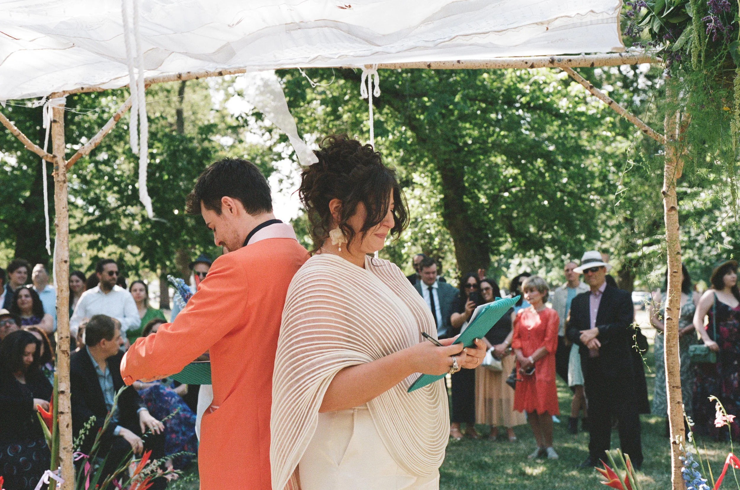 A woman and a man stand back to back under a decorated outdoor canopy during a wedding ceremony, with guests watching in the background.