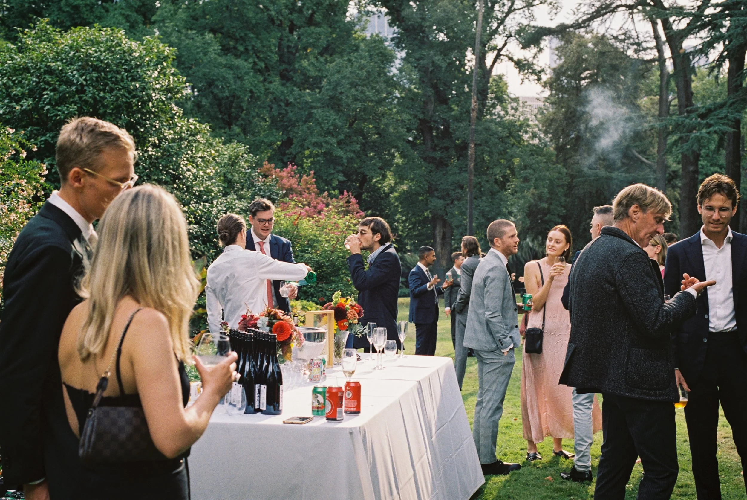 People gathered at an outdoor social event in a garden with tables, drinks, and flowers, enjoying conversations.