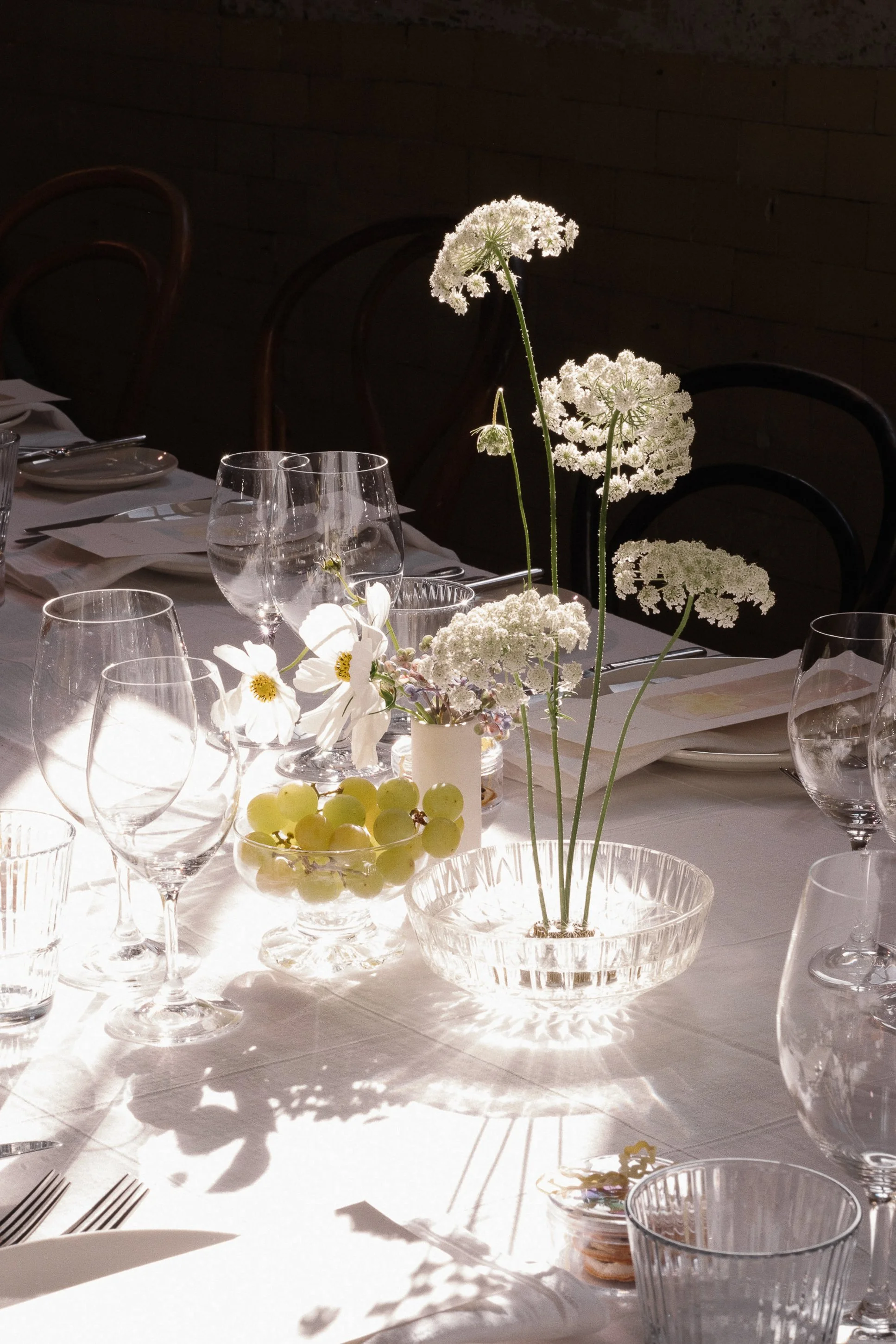 Elegant dining table set with white flowers, grapes, and glassware in sunlight.