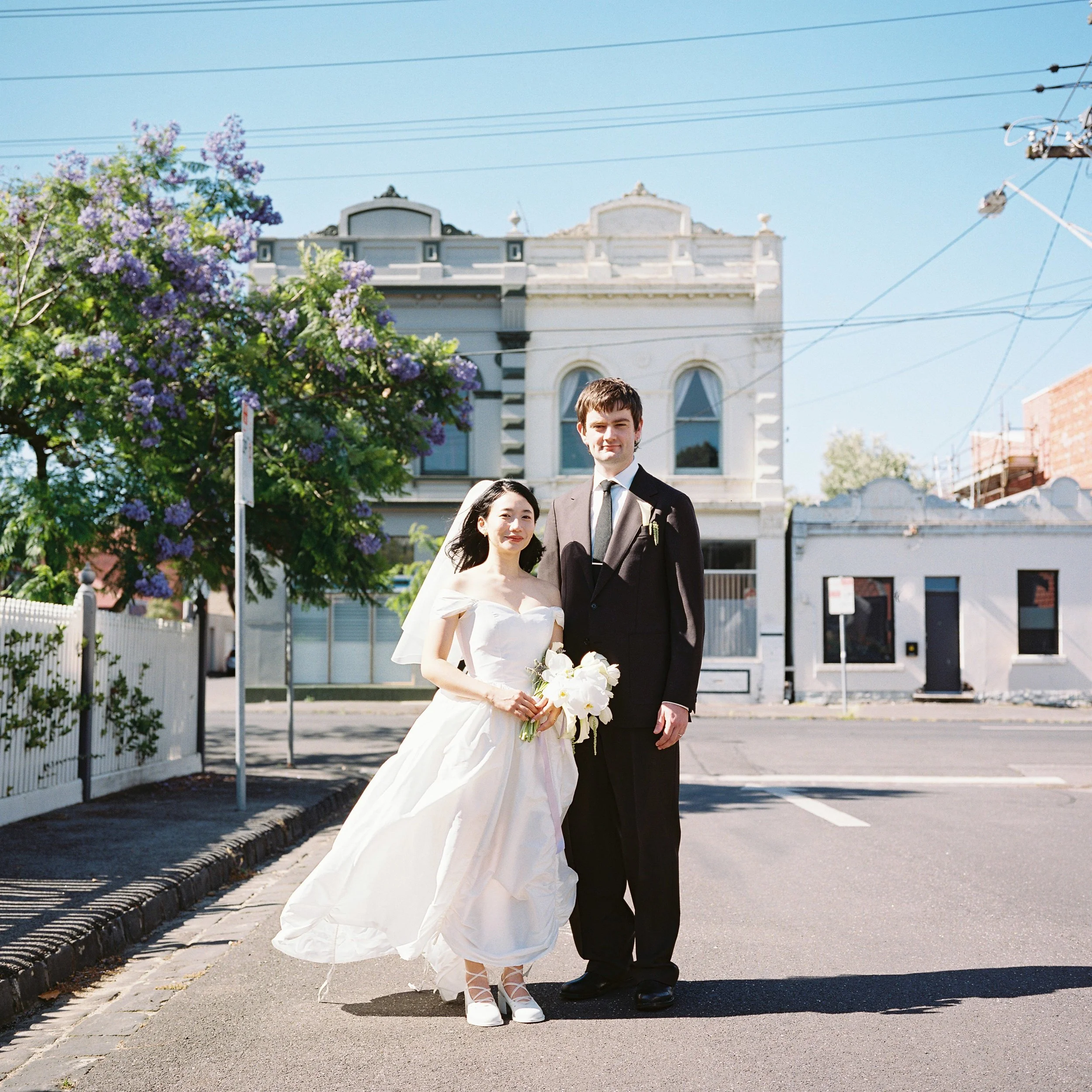 A bride in a white wedding dress holding a bouquet standing next to a groom in a black suit on a city street, with purple flowering tree behind them and old buildings in the background.