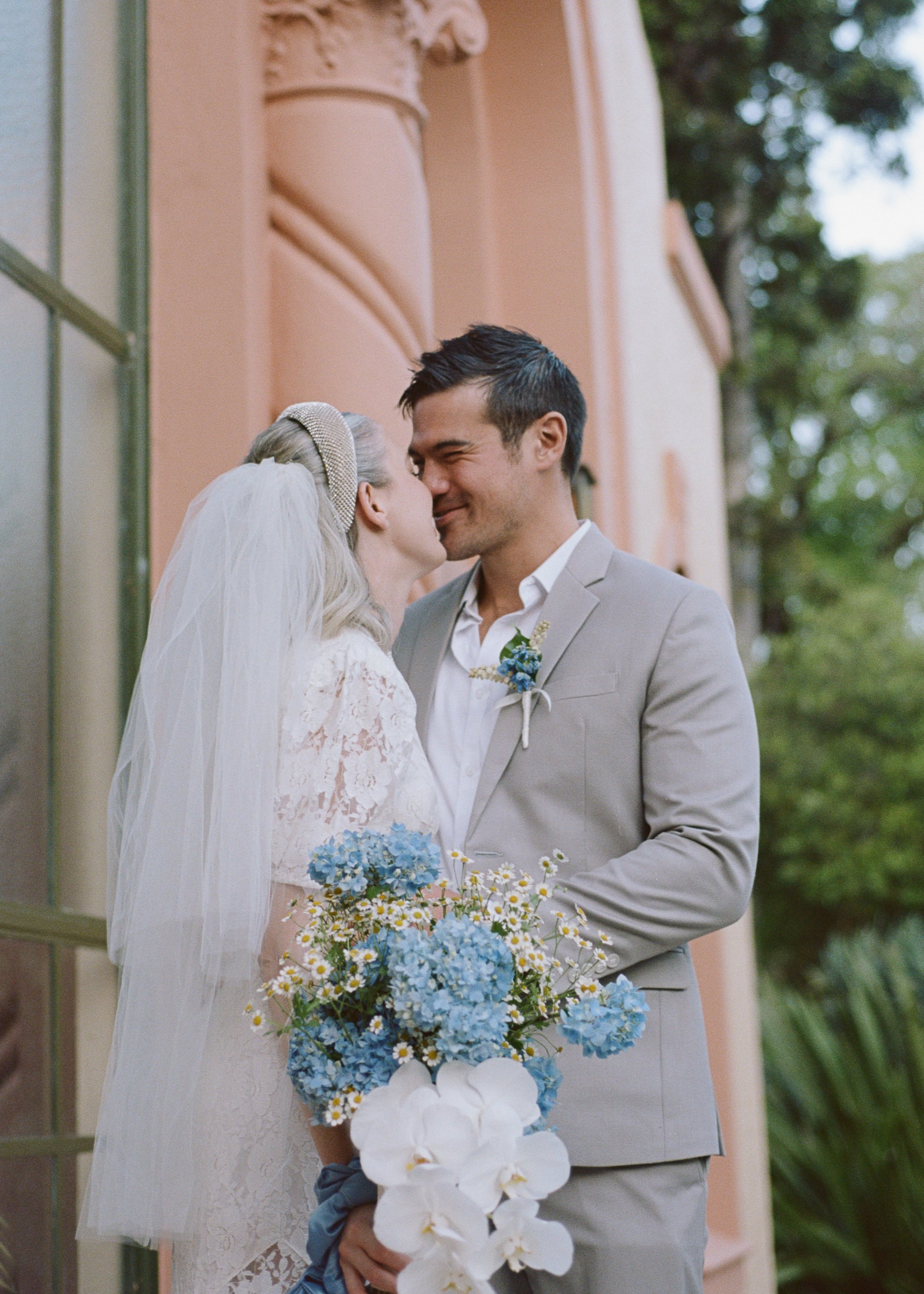 A bride and groom on their wedding day sharing a kiss outside a building with pink walls, surrounded by greenery, with the bride holding a bouquet of blue and white flowers.
