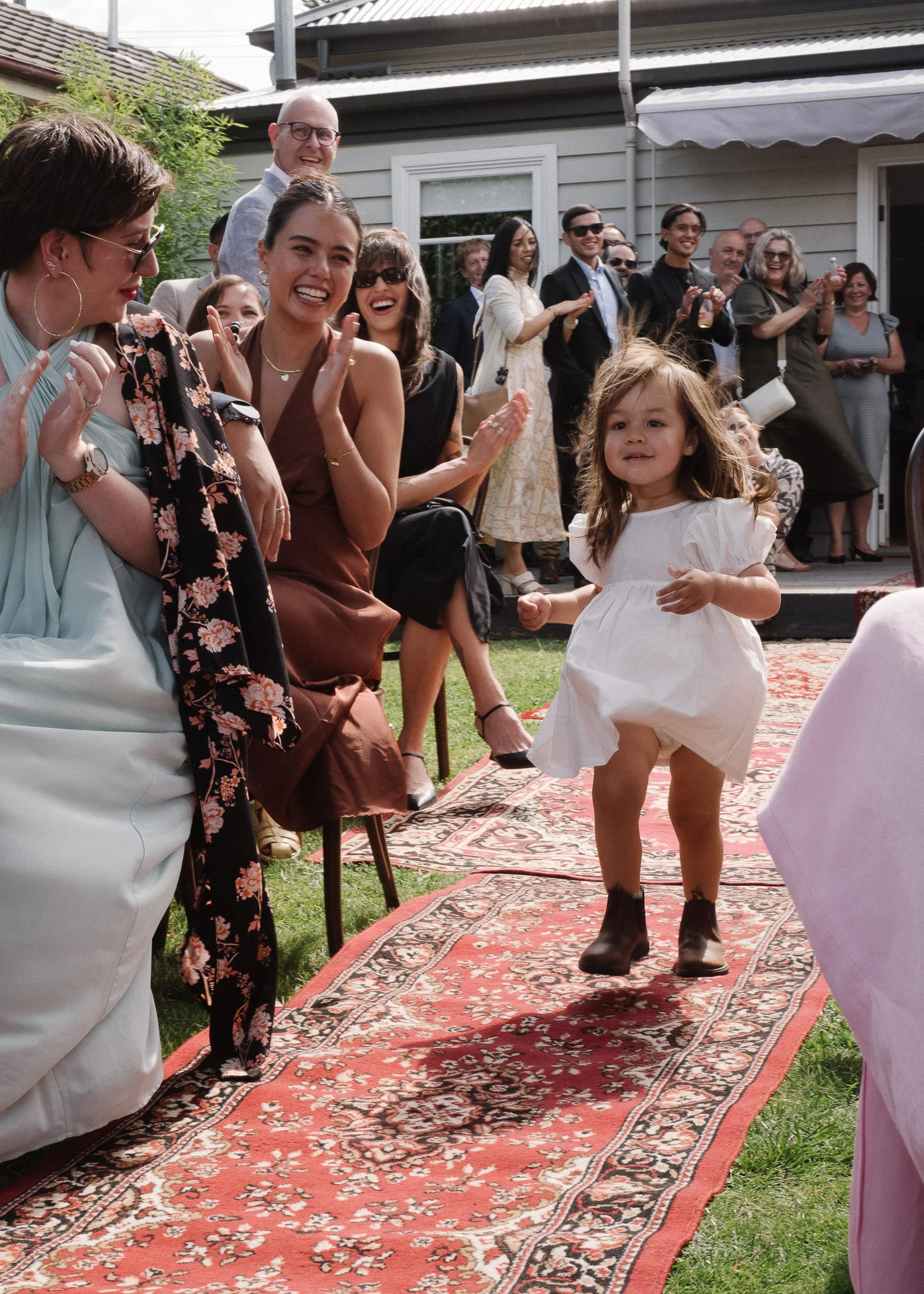 A young girl running down a red patterned rug outdoors at a celebration, with smiling adults sitting and standing, clapping, and watching her.