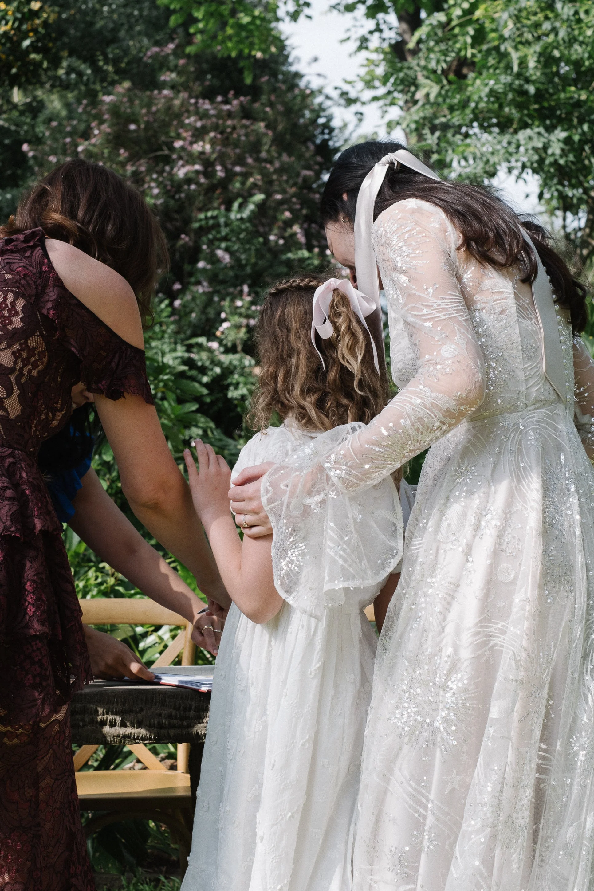 A woman in a white lace wedding dress is holding hands with a young girl in a white dress, during a wedding ceremony outdoors with greenery in the background.