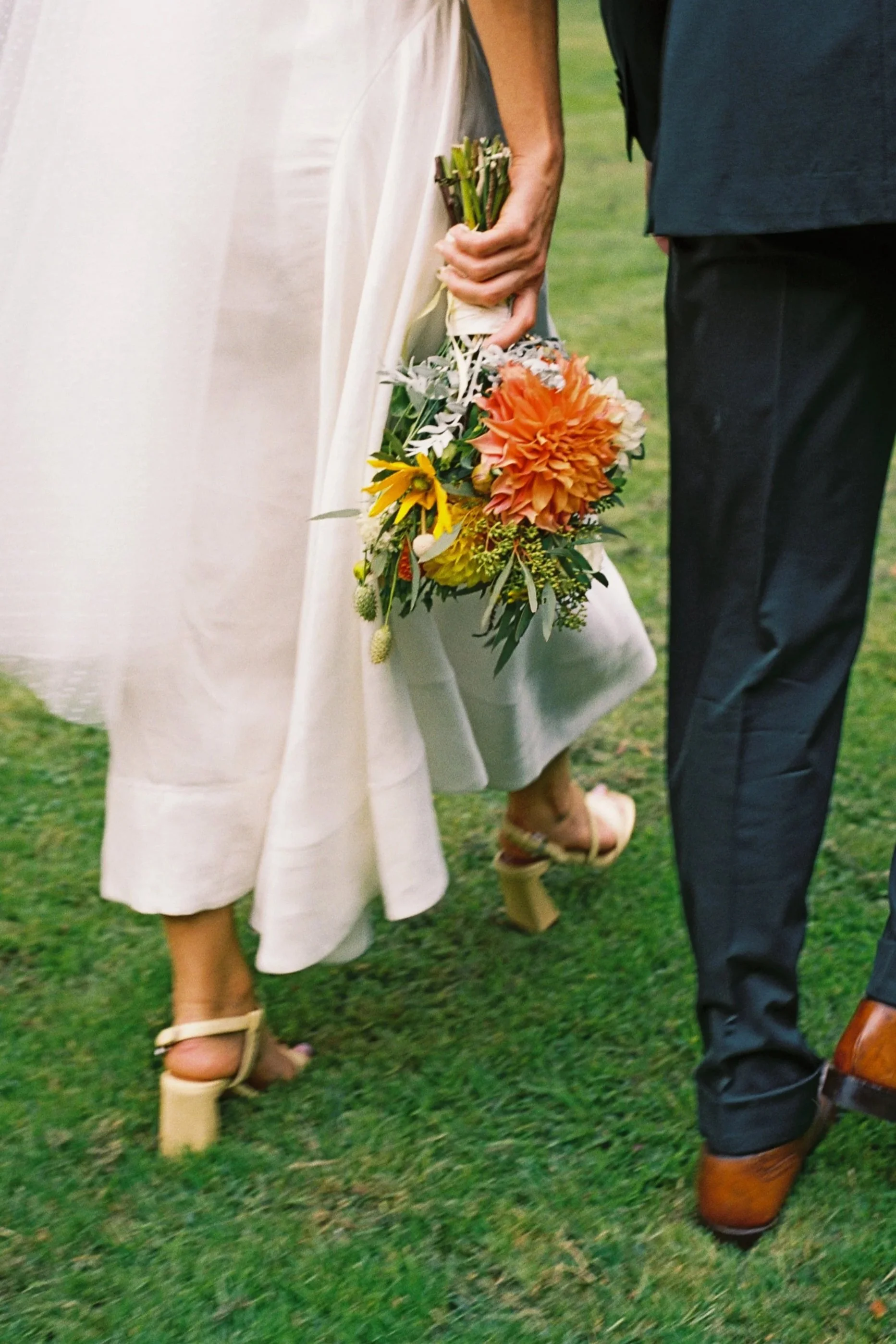 Close-up of a bride and groom holding a wedding bouquet, with only the lower half of their bodies visible on a grassy outdoor setting.