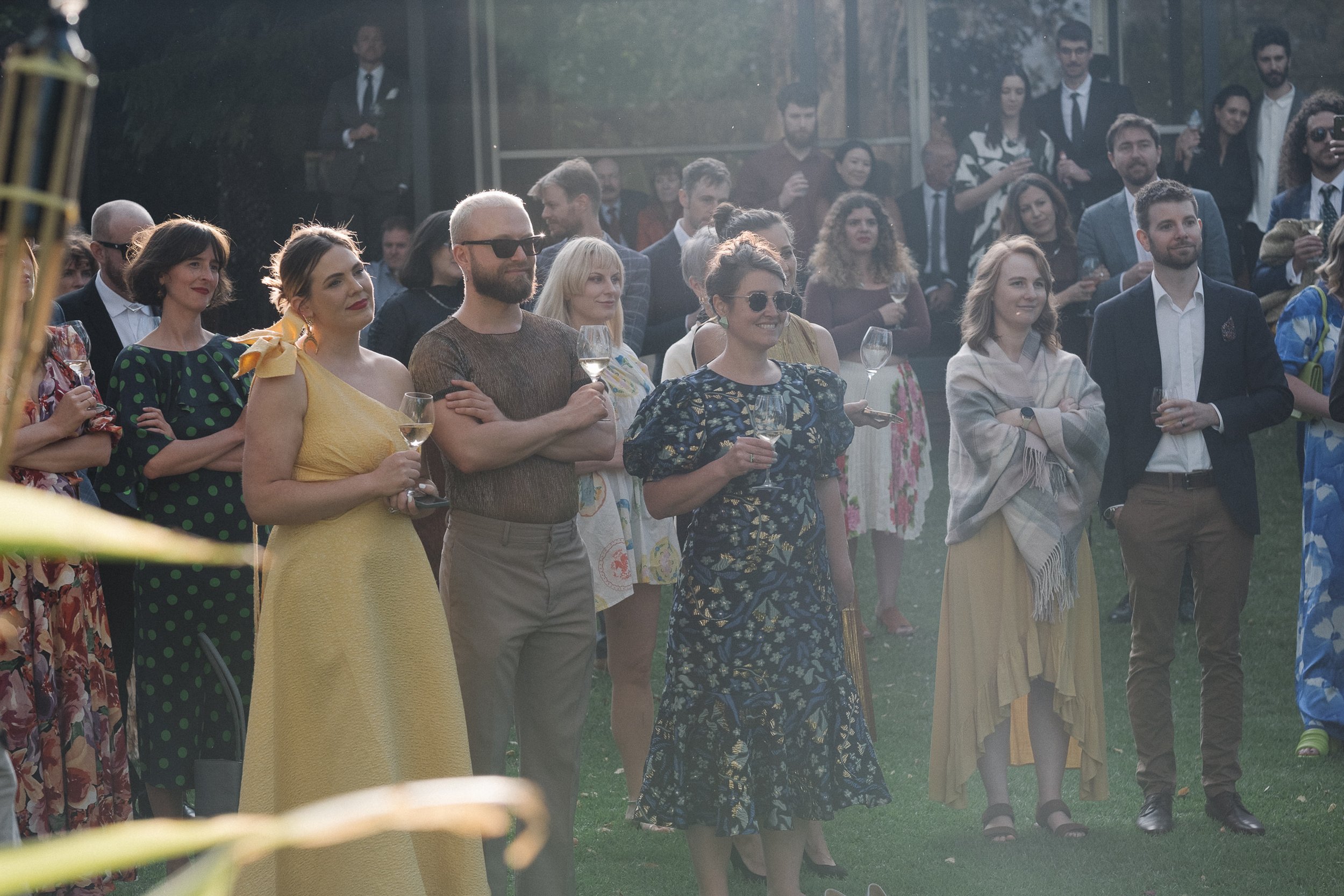 People attending an outdoor social event, some holding wine glasses, dressed in elegant and casual summer attire, with a modern building in the background.
