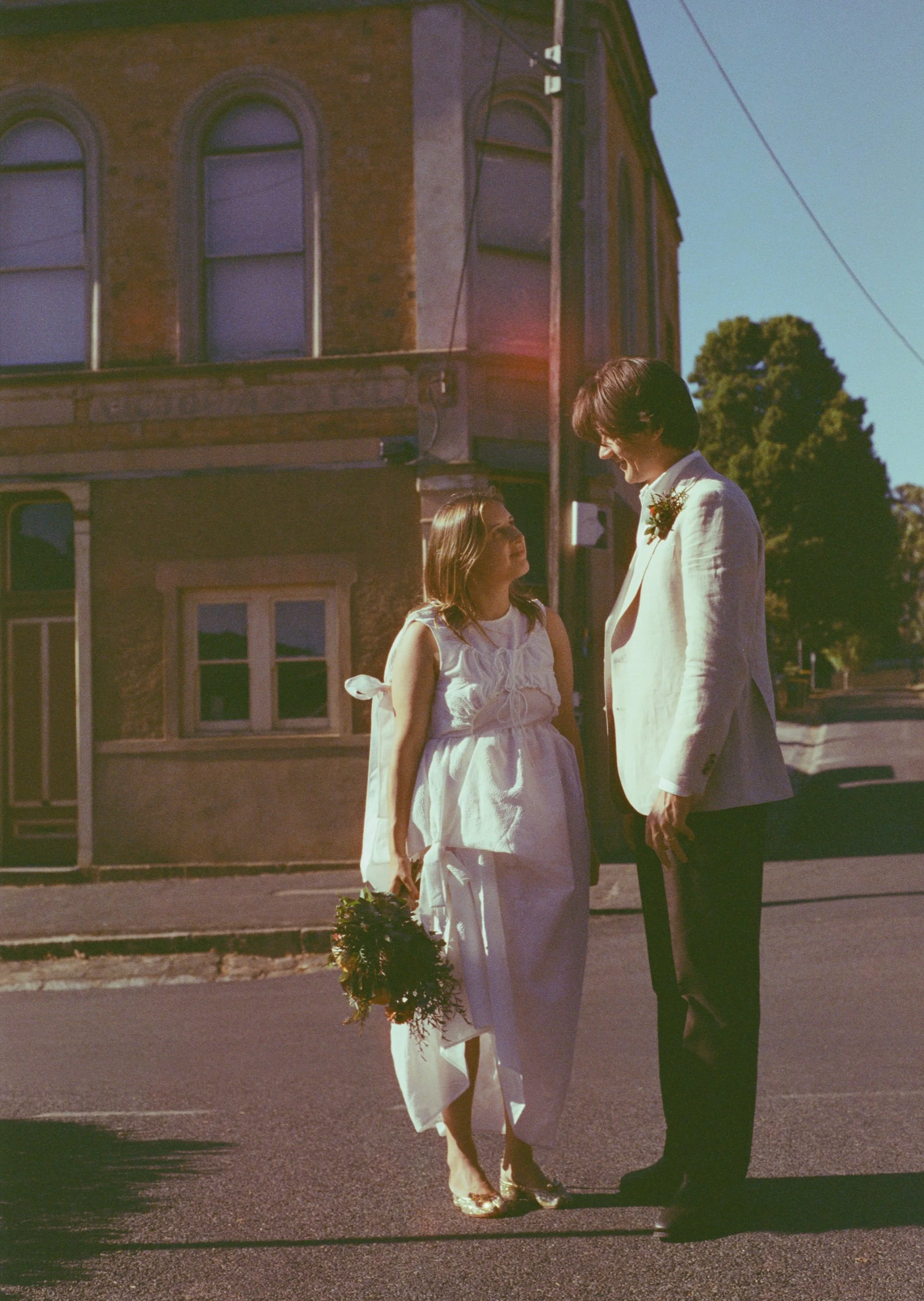 A bride and groom standing on a street during daytime, looking at each other, with a vintage brick building in the background. The bride is holding a bouquet and wearing a white dress, and the groom is dressed in a white suit jacket and dark pants.