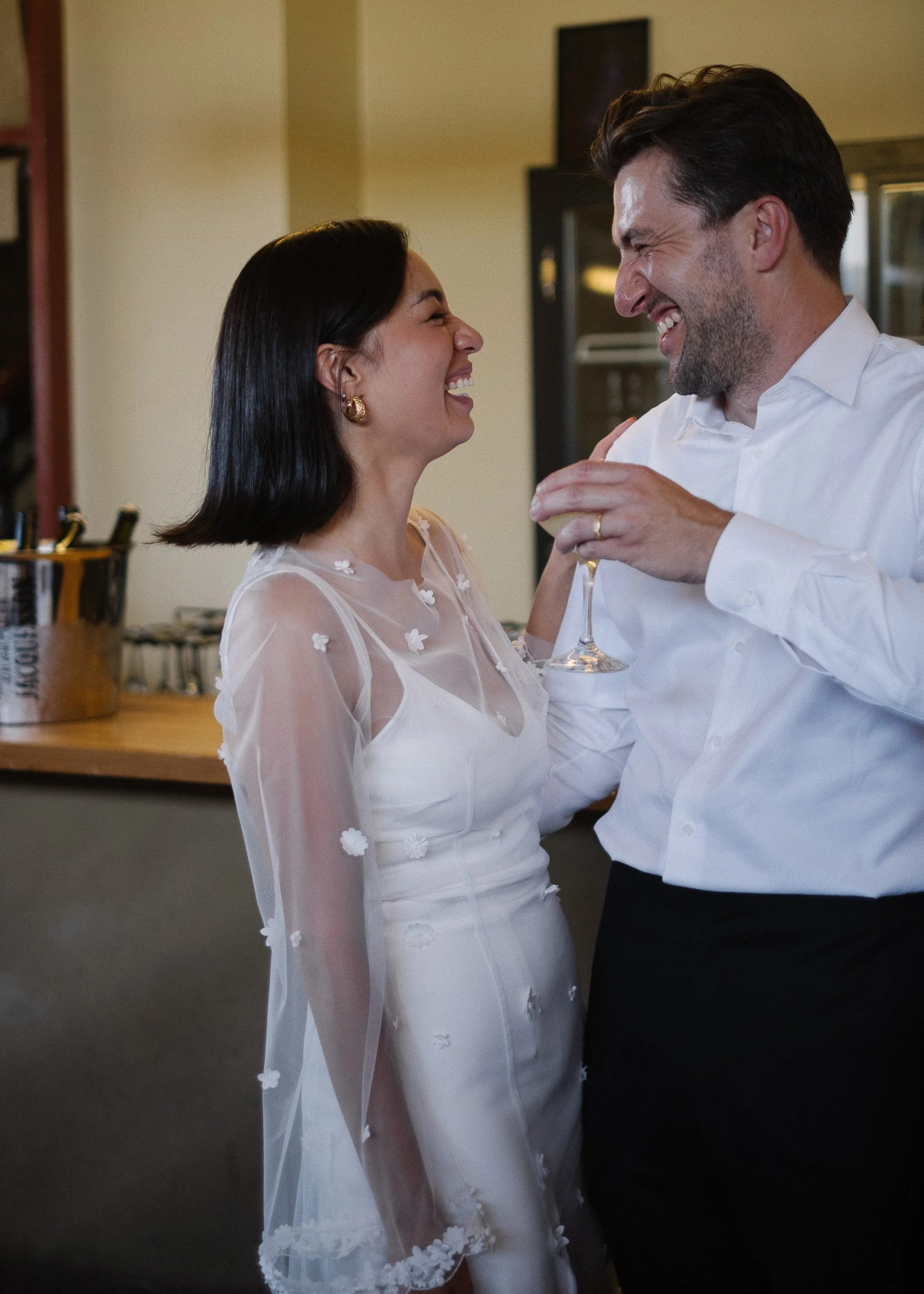 A couple celebrating with big smiles, with the man holding a glass of wine. They are dressed in elegant attire, and the woman is wearing a white dress with floral details.