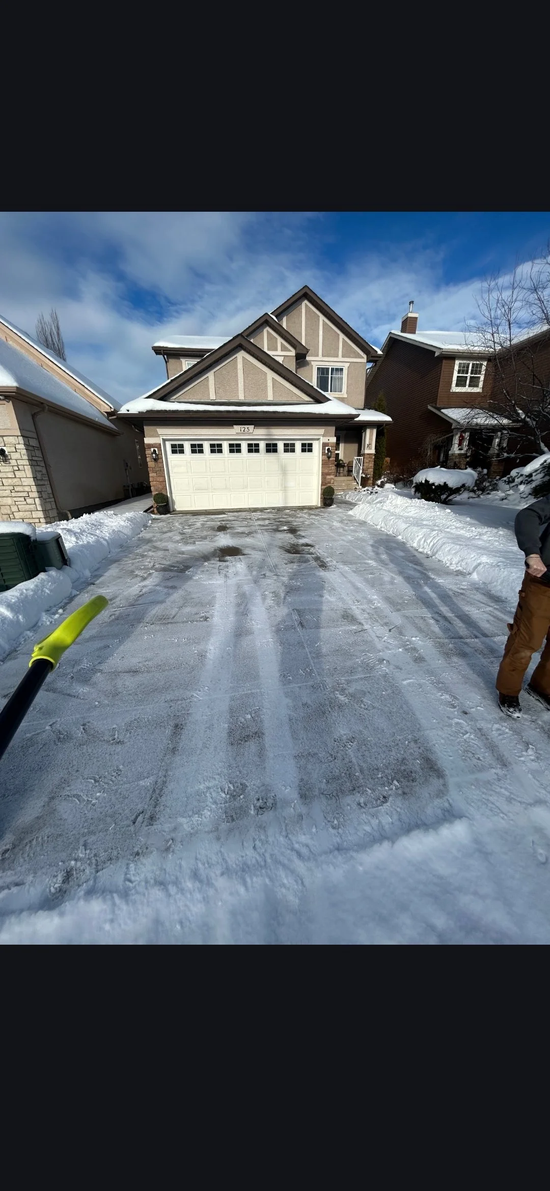 Snow-covered driveway leading to a house with a closed garage door, with a person standing on the right side and snow on the roofs and surrounding areas.