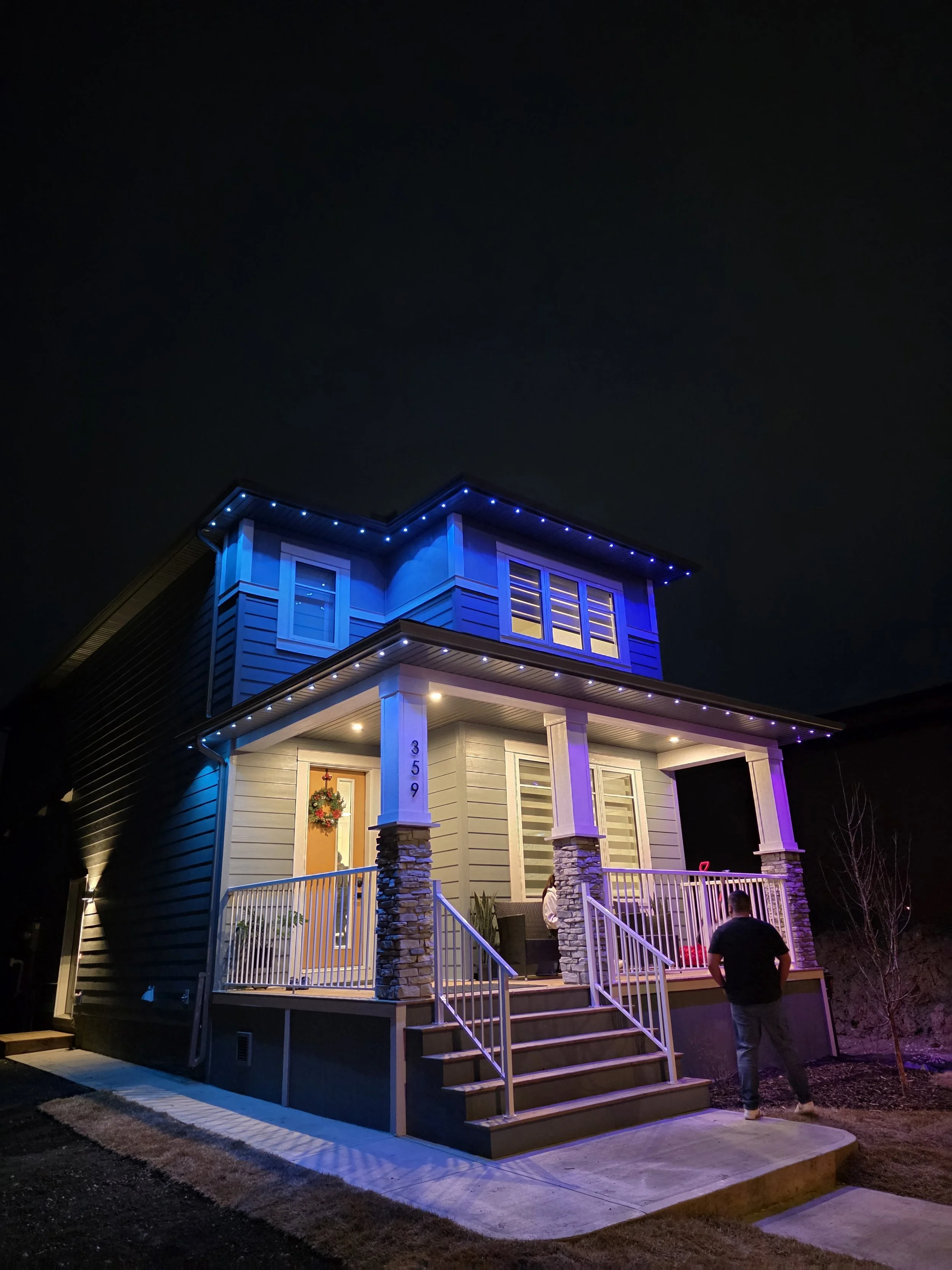 A two-story house decorated with blue and white string lights outside at night. The house has a porch with stairs, stone pillars, and a man standing on the sidewalk.