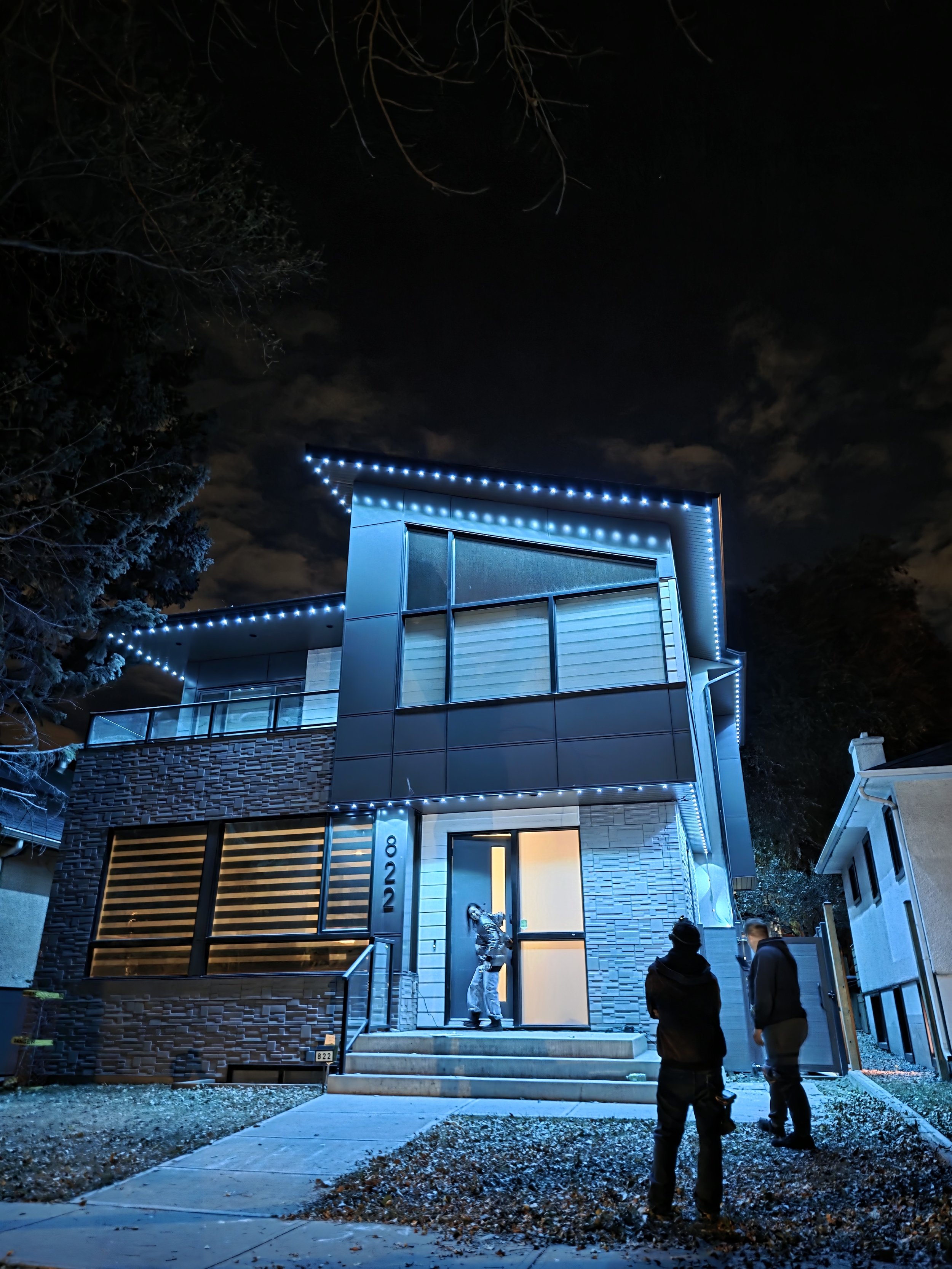 Nighttime exterior of a modern, two-story house decorated with blue LED lights outlining the roof and windows. There are three people standing and talking outside the house near the front steps.