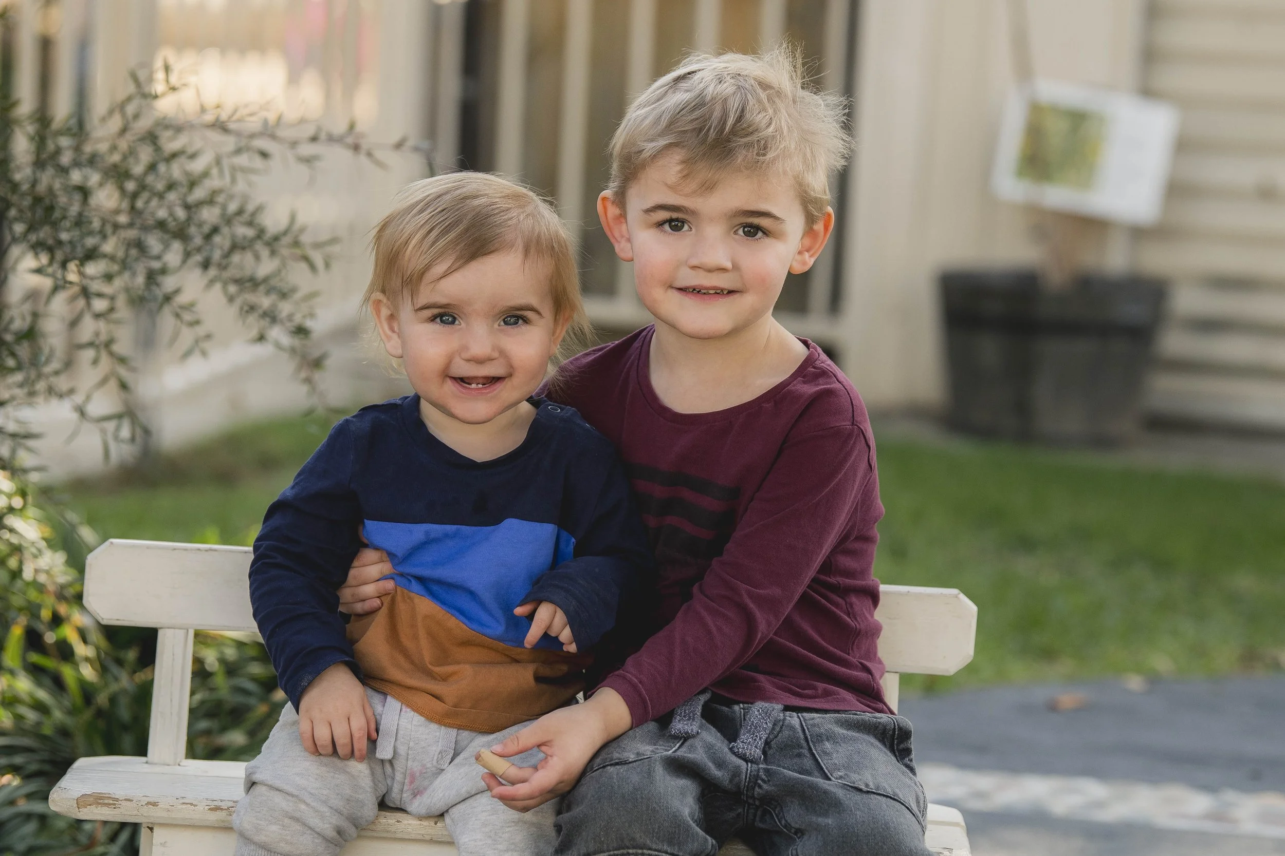 Two young children, a boy and a girl, sitting on a white park bench outdoors with a blurred background of greenery and building walls. The girl has light brown hair and is wearing a navy blue and brown shirt, smiling with her arm around the boy. The boy has light blonde hair, wears a maroon shirt, and is smiling facing the camera, with his arm around the girl.