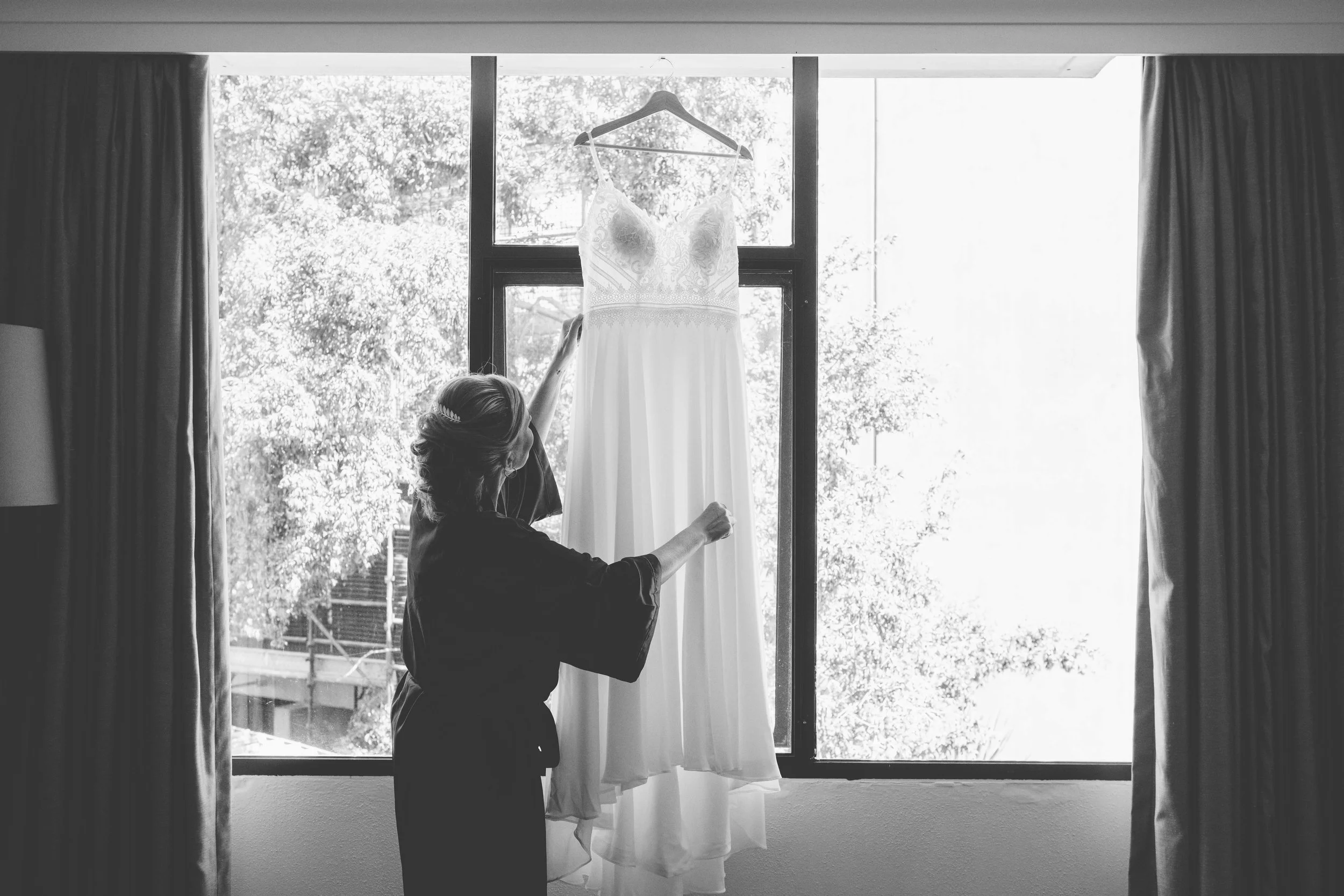 Woman hanging a wedding dress in front of a large window with curtains on each side.