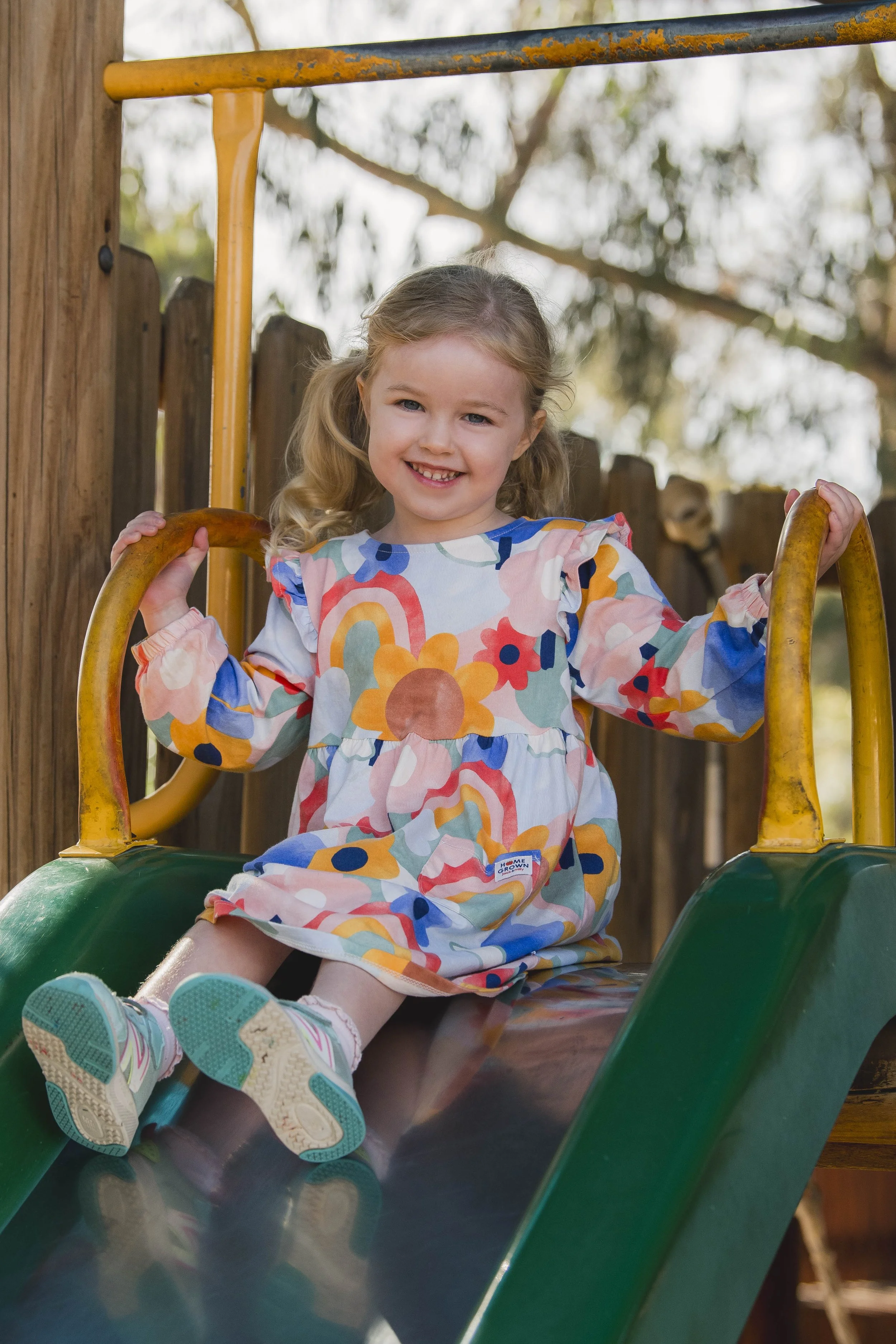 A young girl with curly blonde hair and a big smile sitting at the top of a green slide on a playground, holding onto orange safety bars on either side, wearing a colorful dress with floral patterns, white socks, and sneakers, with trees in the background.