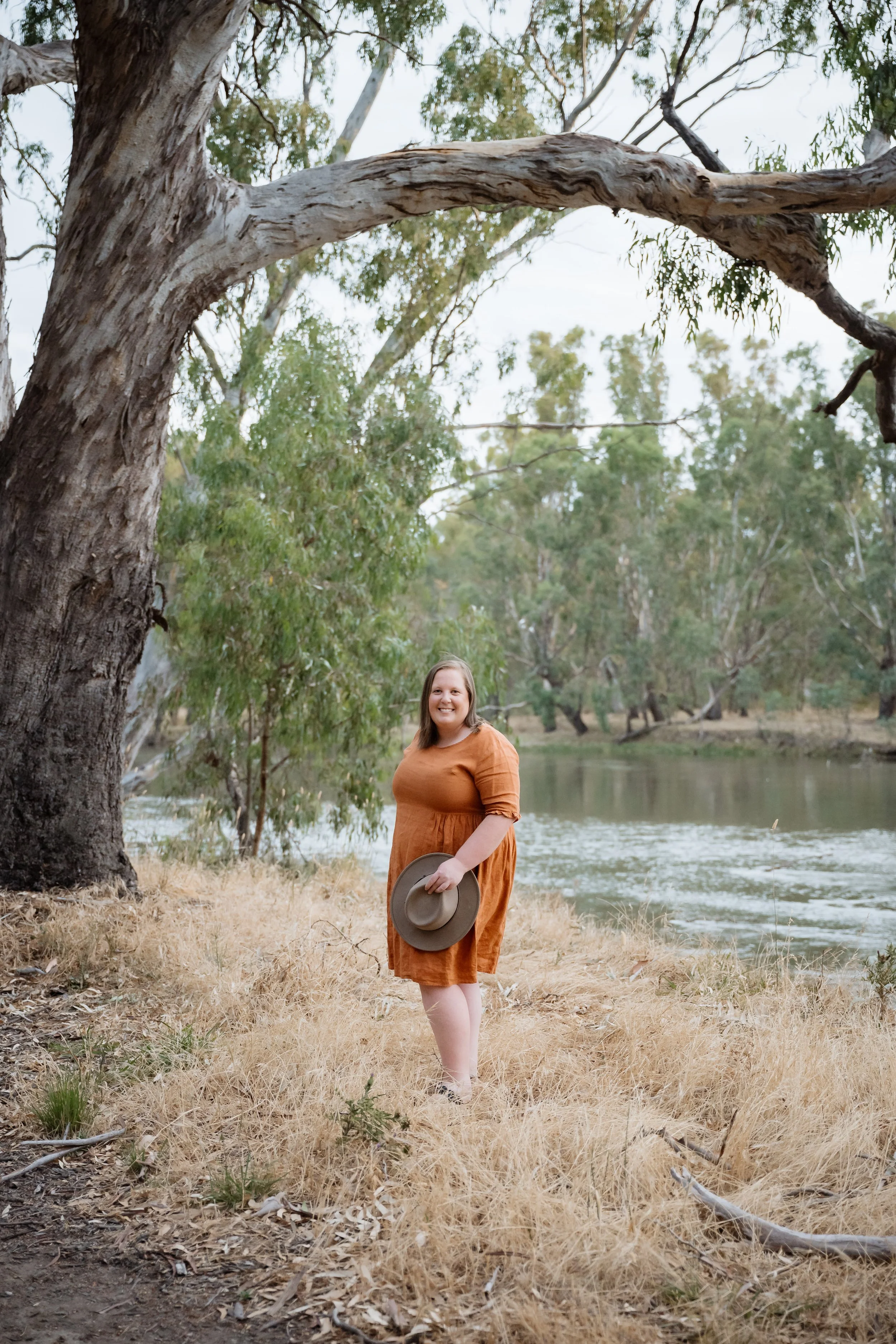 A woman in an orange dress holding a wide-brimmed hat stands on dry grass near a river, with tall trees and a large, curved tree branch overhead, during daytime.