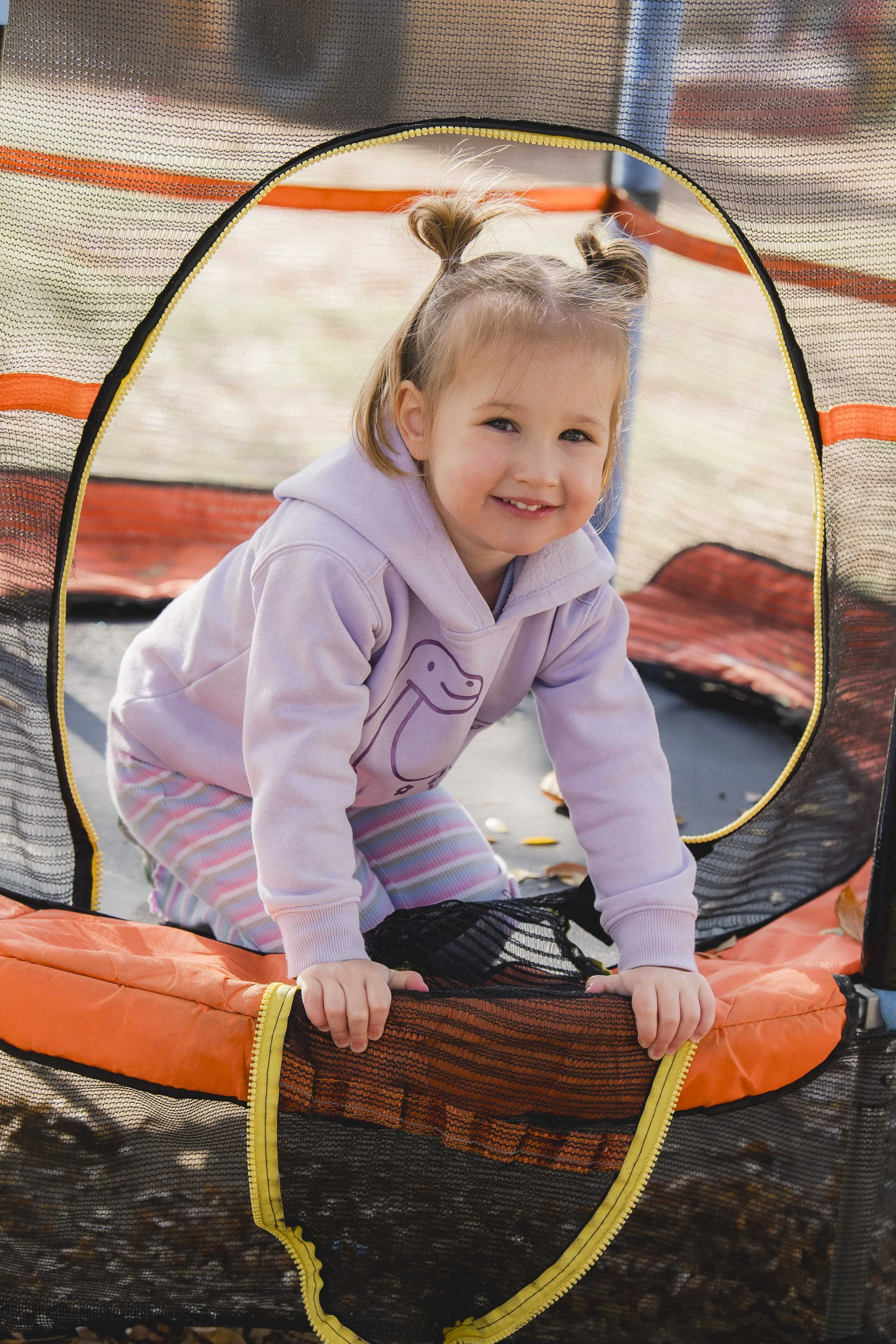 A young girl with two pigtails climbing out of an orange and black zippered play tent, smiling with her hands on the ground, outdoors surrounded by fallen autumn leaves.