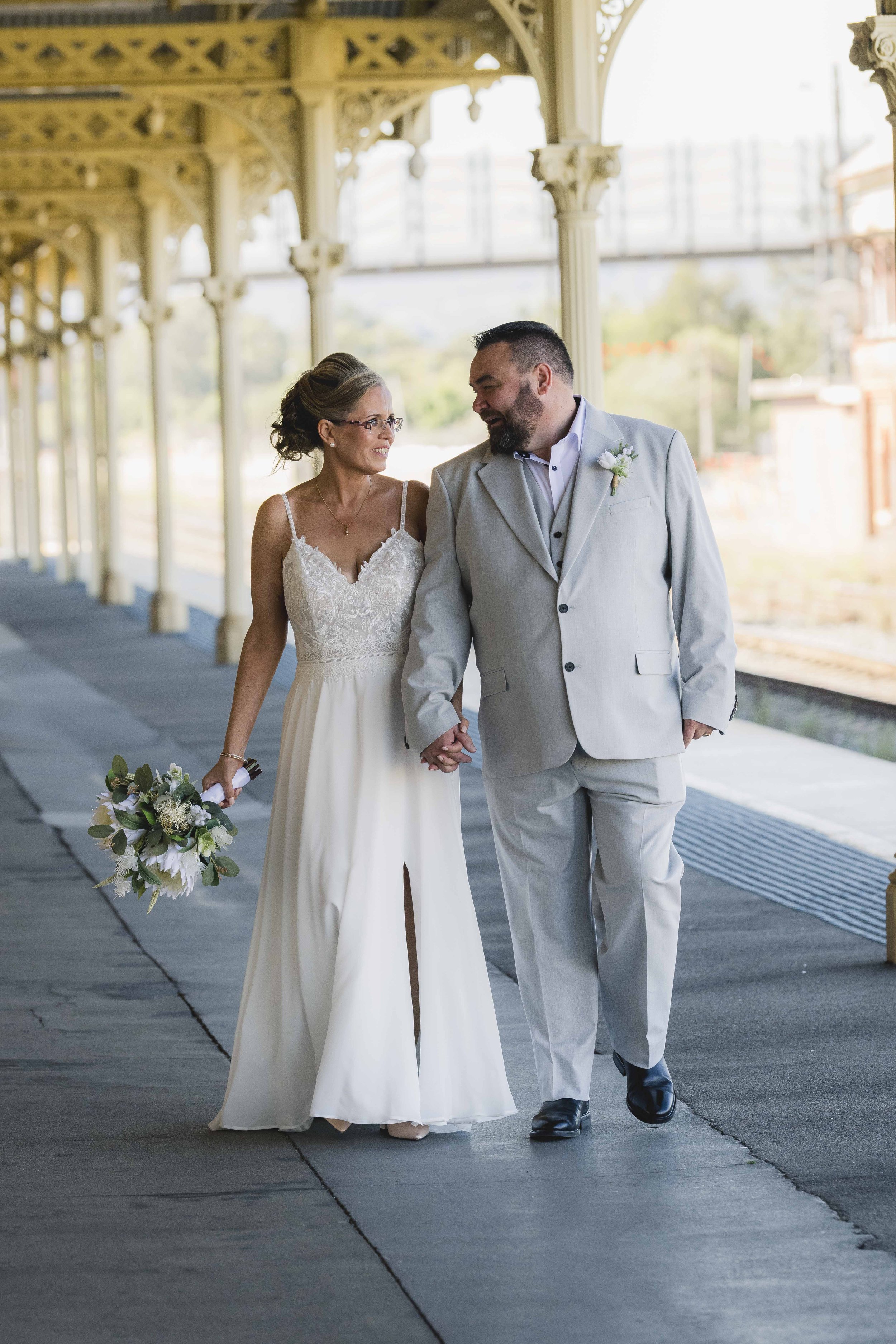 A married couple walking hand in hand under a decorative pavilion during their wedding. The bride is wearing a white dress and glasses, carrying a bouquet, while the groom is dressed in a light gray suit with a boutonniere.