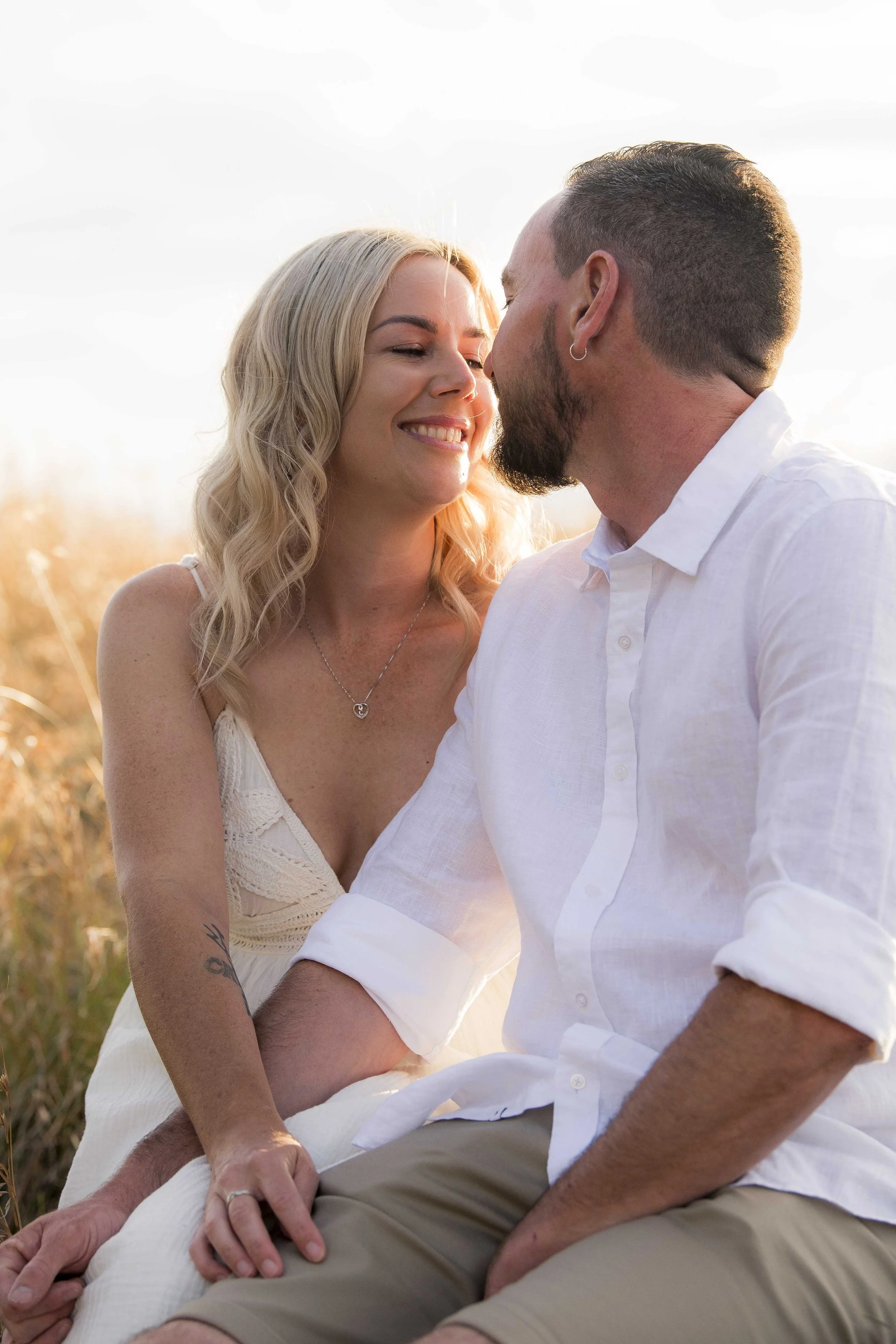 A smiling woman with blonde hair and a man with short hair and a beard sit closely together outdoors in a field during sunset, sharing an intimate moment.