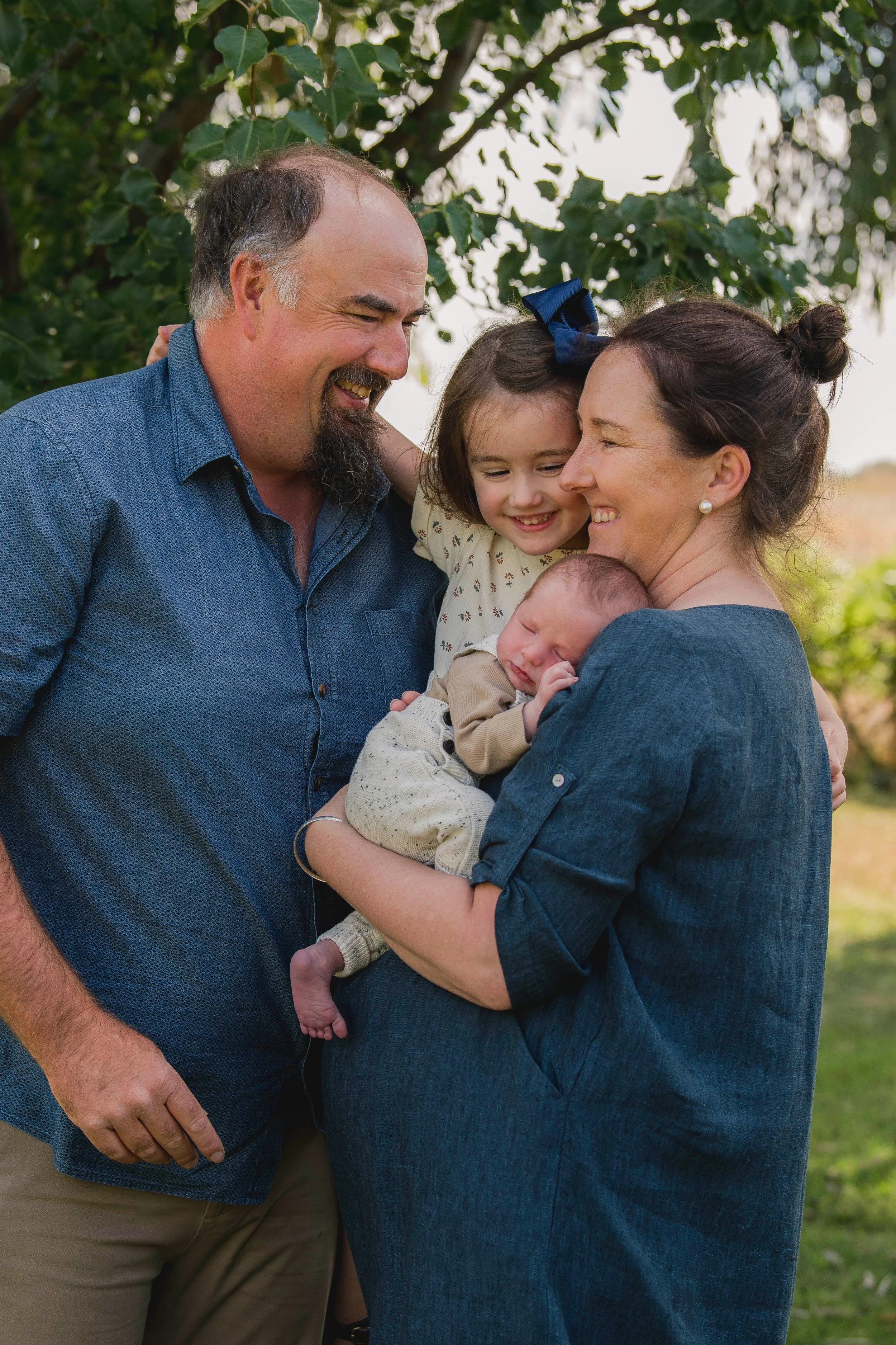 A happy family outdoors, with a man, woman, and two children, sharing a joyful moment together in front of green trees.