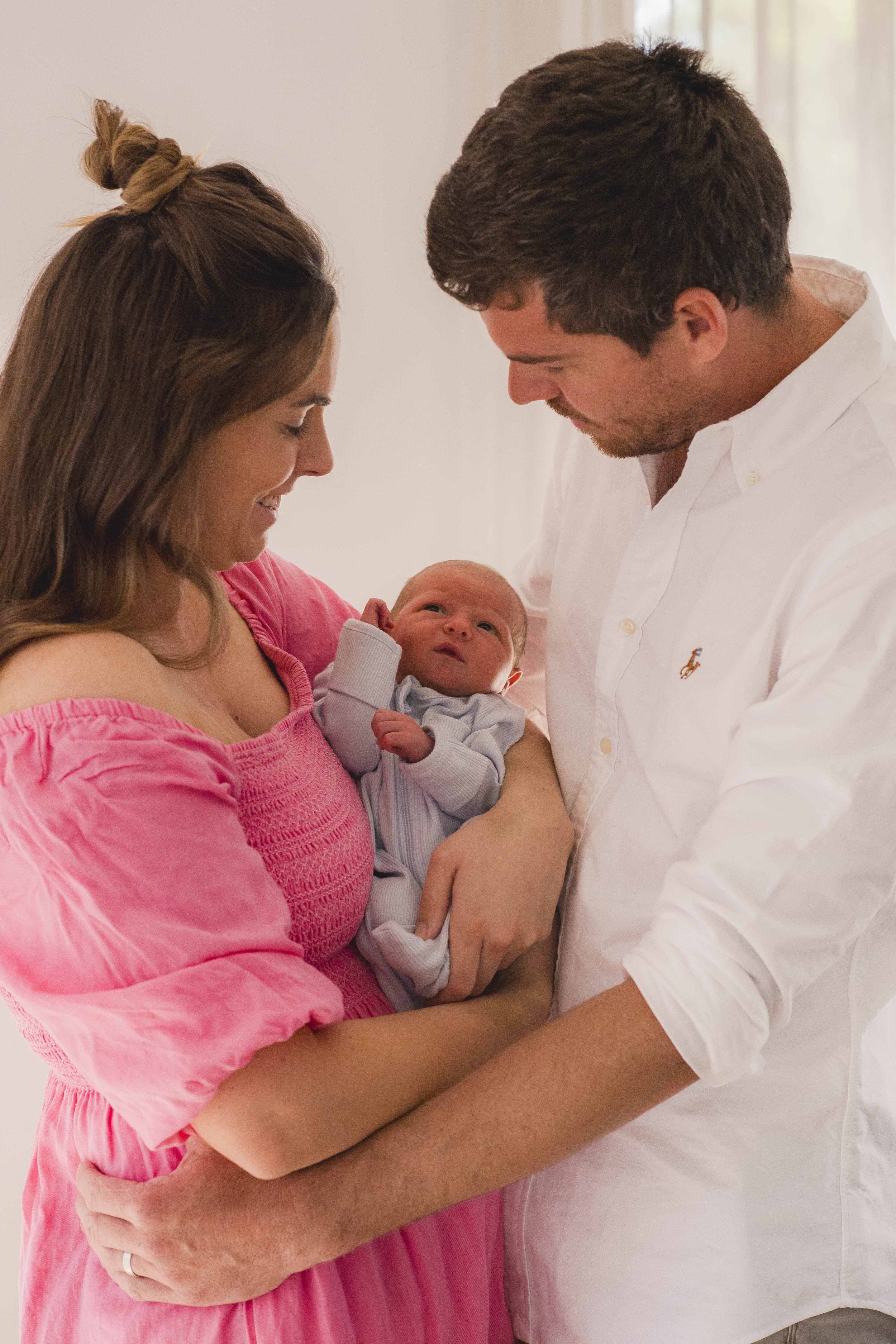A woman in a pink dress holds a newborn baby, while a man in a white shirt looks at the baby, all standing close together and smiling.