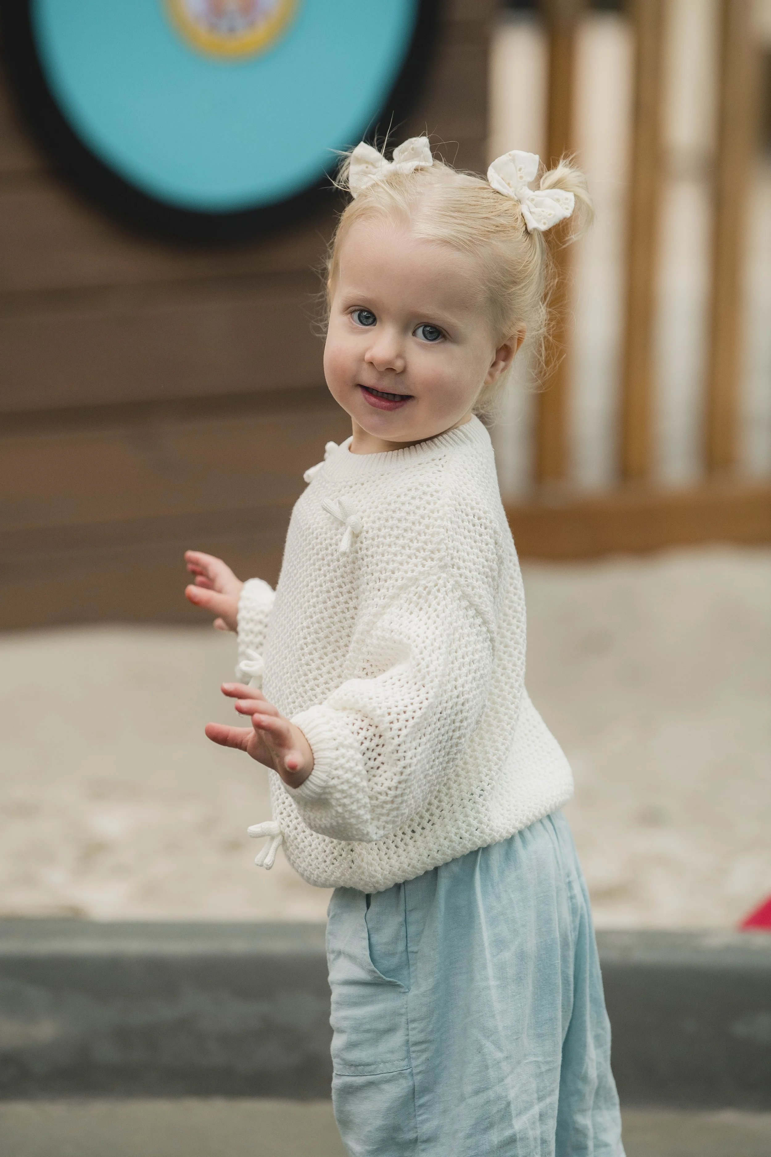 A young girl with blonde hair tied in two ponytails with white bows, wearing a white knitted sweater and light blue pants, standing outdoors on a street with a blurred background.