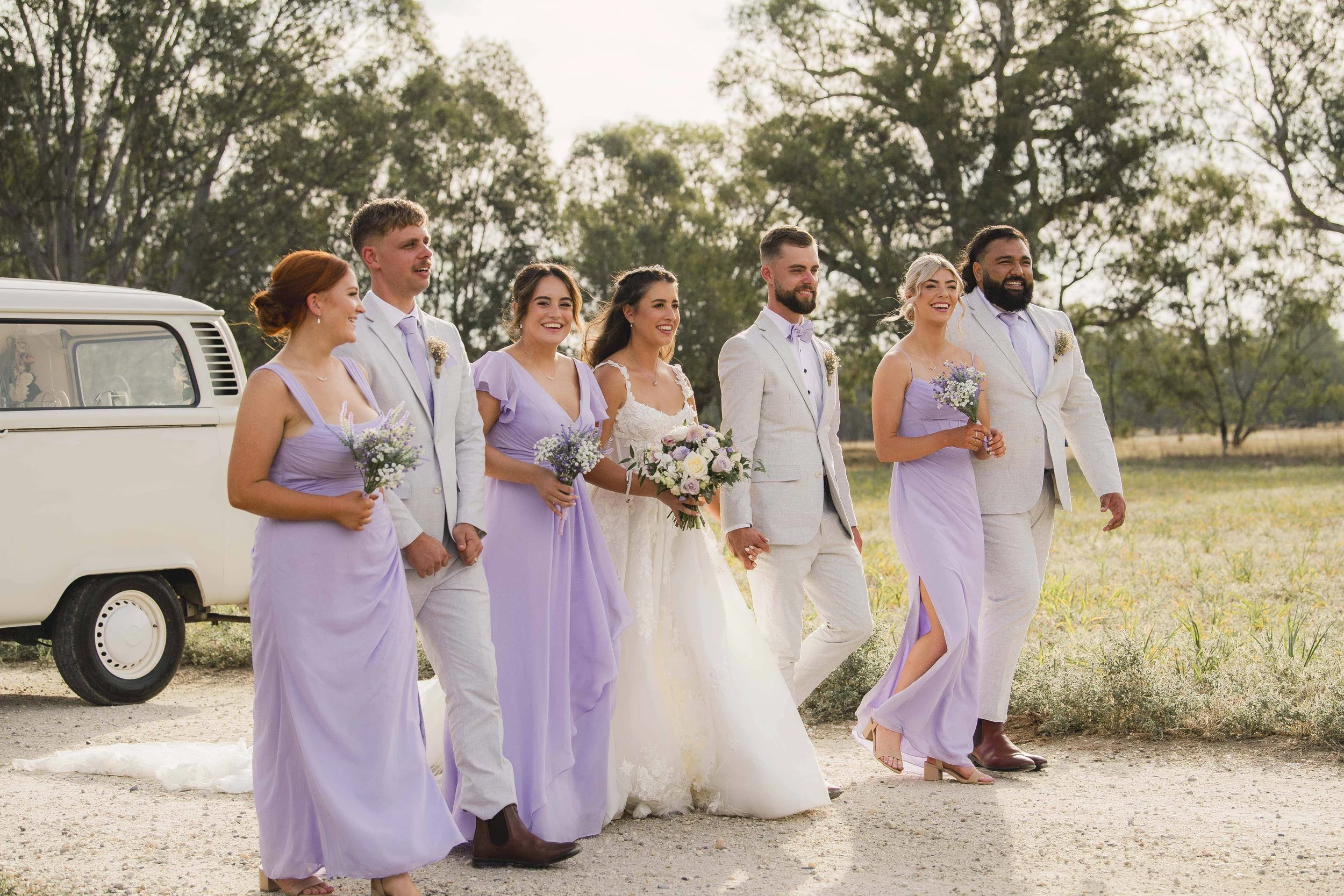 A wedding party dressed in lavender and white, walking outdoors in a field with trees in the background. The bride is in a white wedding dress holding a bouquet, flanked by bridesmaids and groomsmen. The group appears joyful and celebratory.