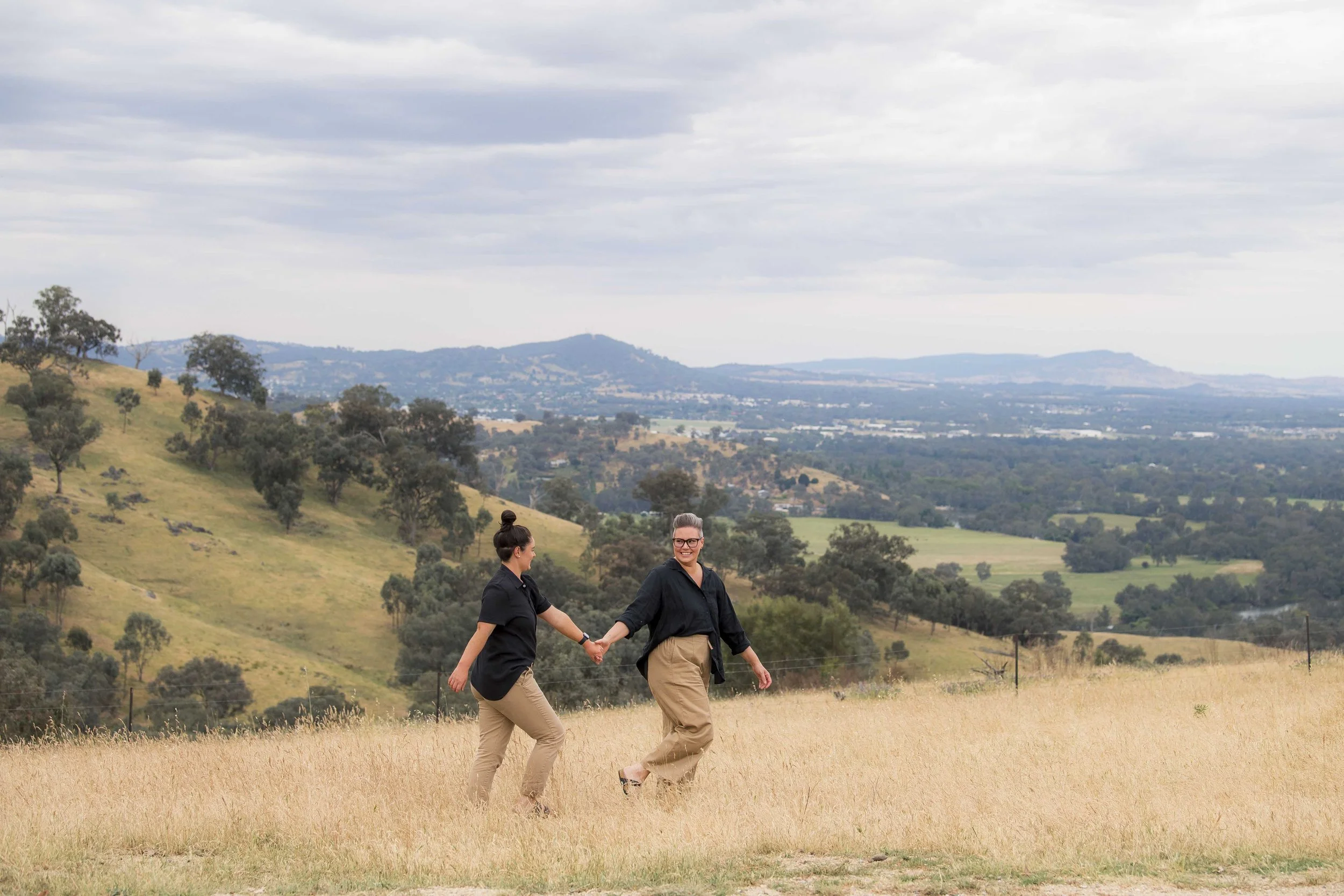 Two people holding hands and walking through a grassy field with rolling hills and trees in the background under a cloudy sky.