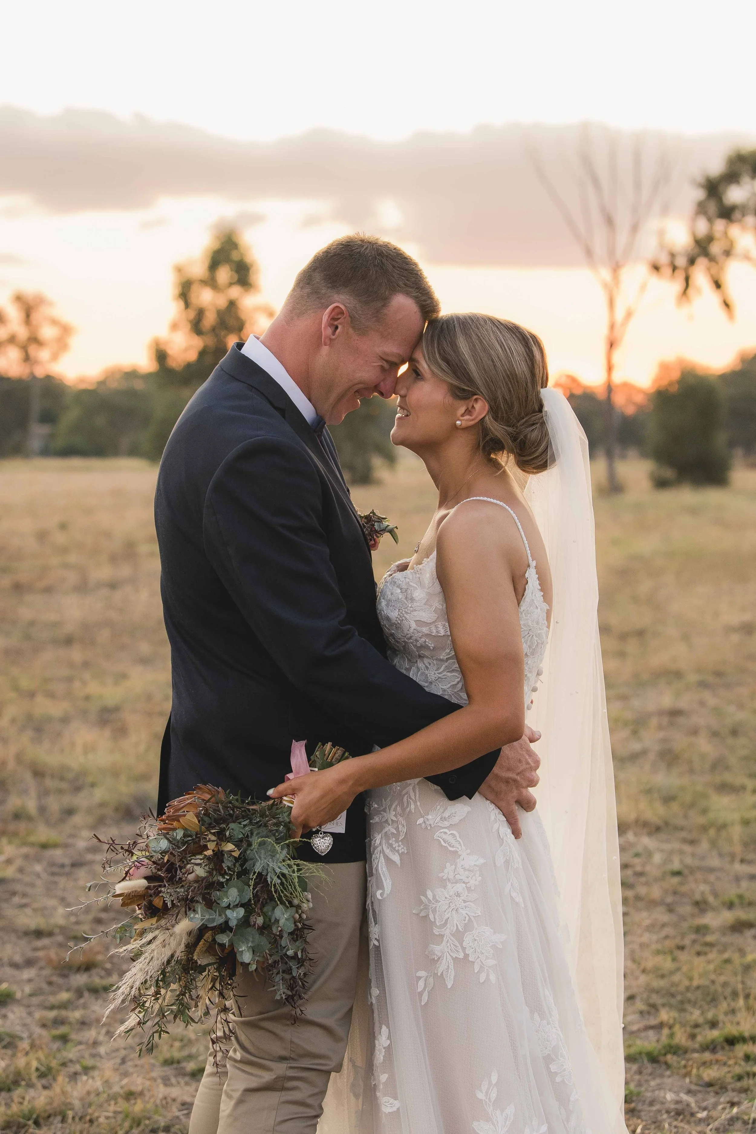 A happy couple on their wedding day standing outdoors at sunset, touching foreheads and holding hands. The groom is wearing a dark suit, and the bride in a white lace dress with a veil. The bride is holding a bouquet of flowers. The background shows trees and a colorful sky.