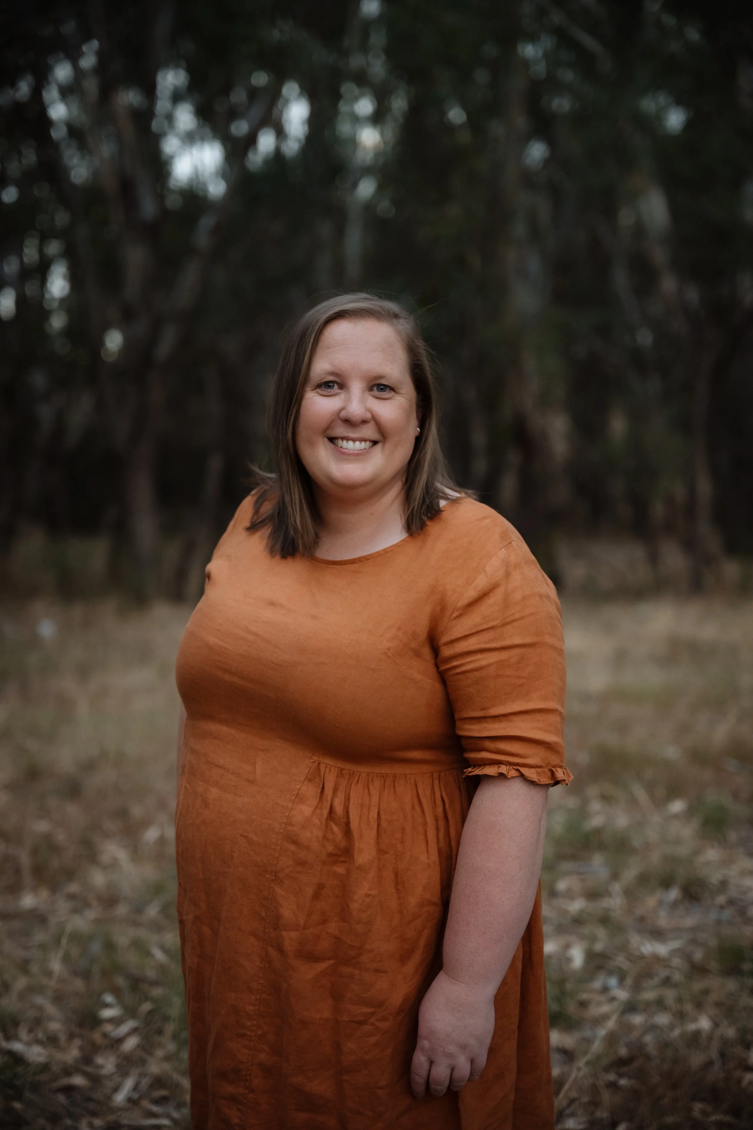 A woman with shoulder-length brown hair smiling outdoors, wearing an orange dress with ruffled sleeves, standing in a natural, wooded setting with blurred trees in the background.