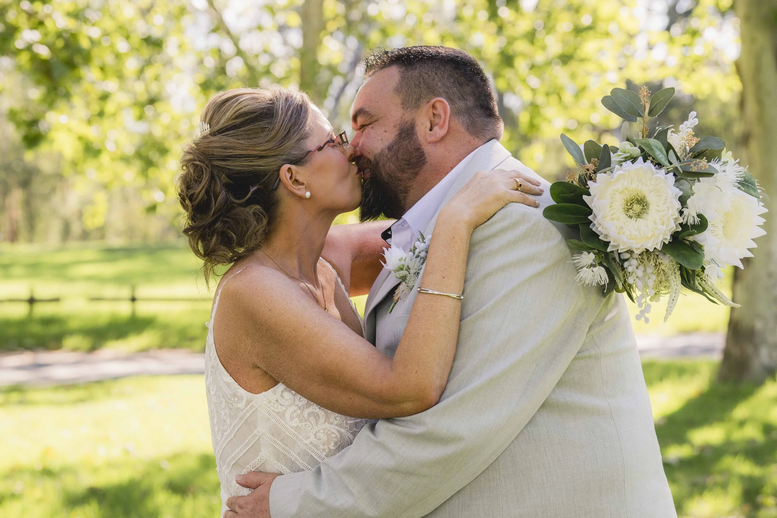 A couple, dressed in wedding attire, sharing a kiss outdoors with green trees in the background. The woman is holding a bouquet of white flowers and wearing glasses, and the man has a beard and short hair.