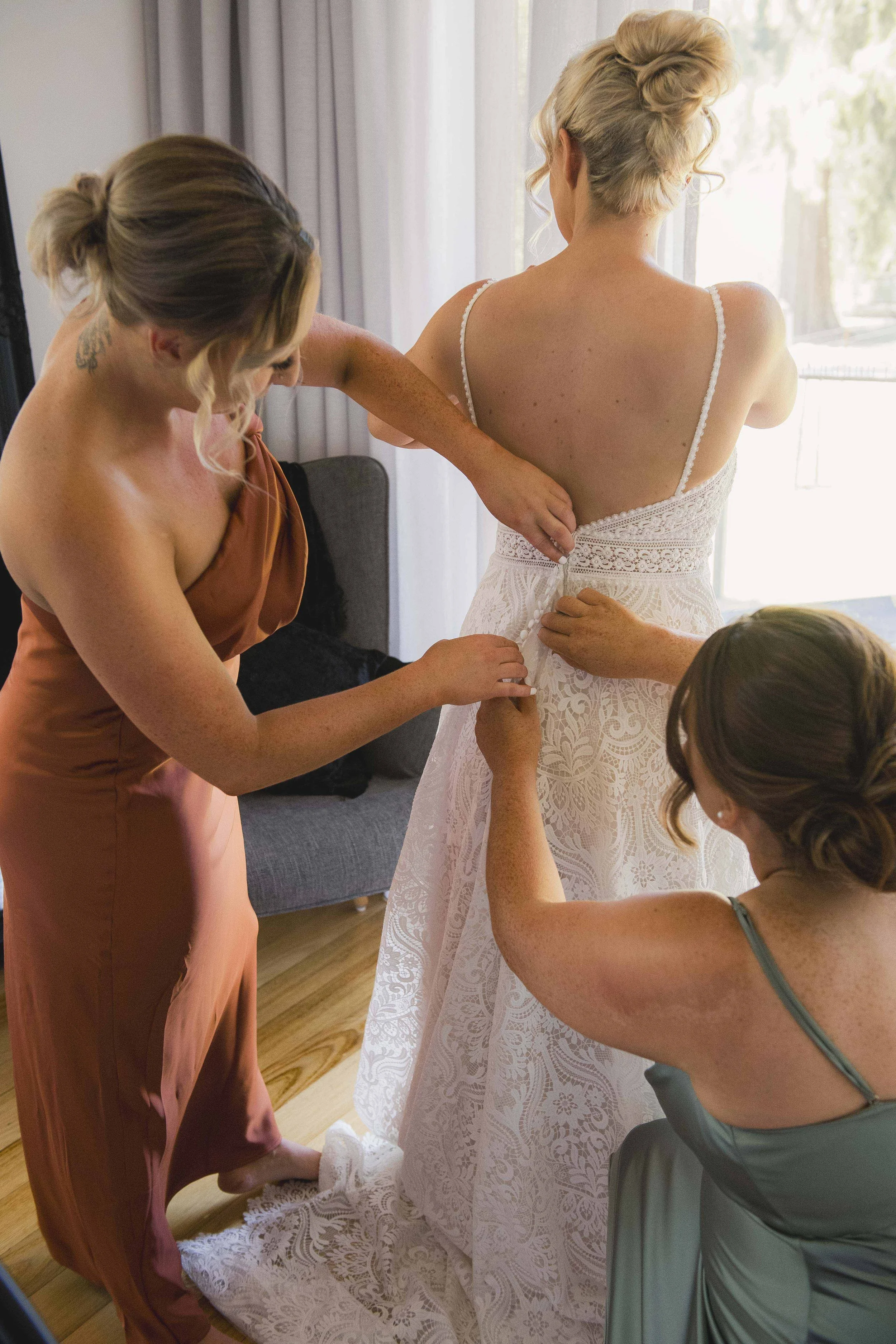Women assisting bride with dress on, in a bright room with large windows and curtains.