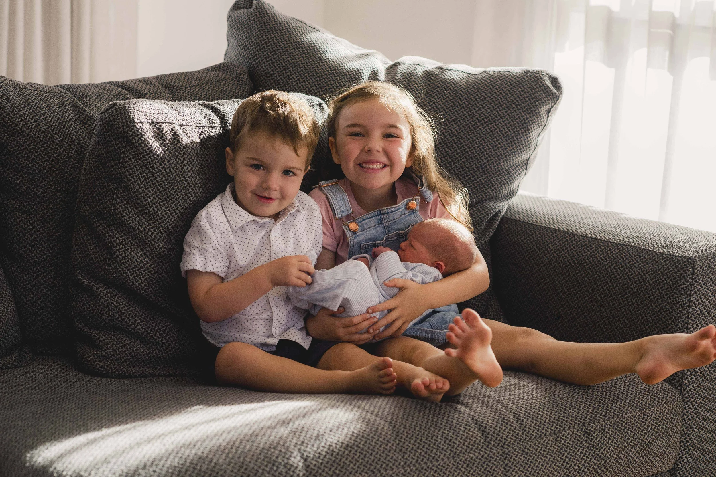 Two young children, a boy and a girl, sitting on a gray couch with a newborn baby. The girl is smiling broadly, and the boy looks calm. Sunlight filters through sheer curtains in the background.