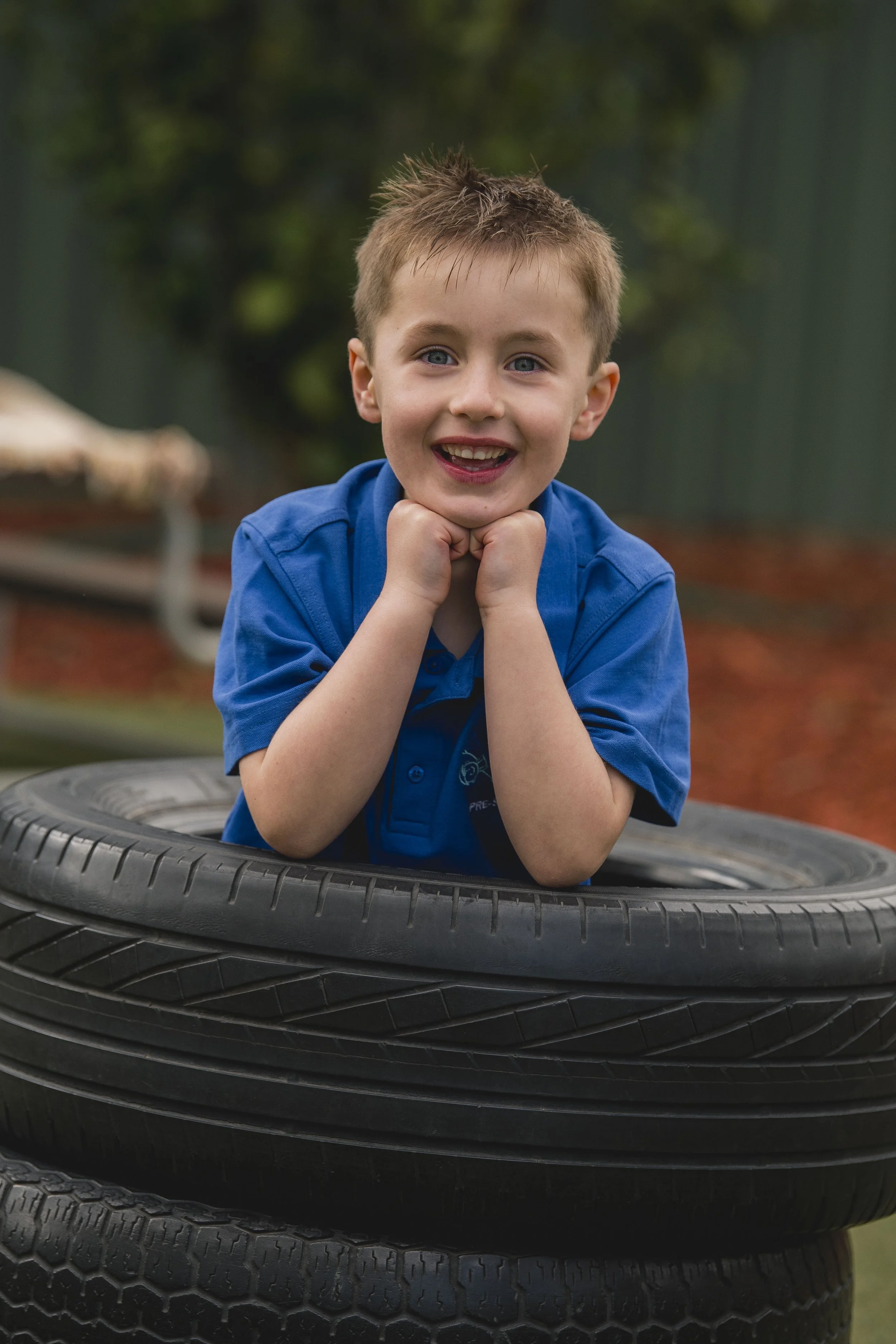 A young boy with short brown hair and blue eyes, wearing a blue shirt, is smiling and resting his chin on his hands inside a stack of black tires outdoors.