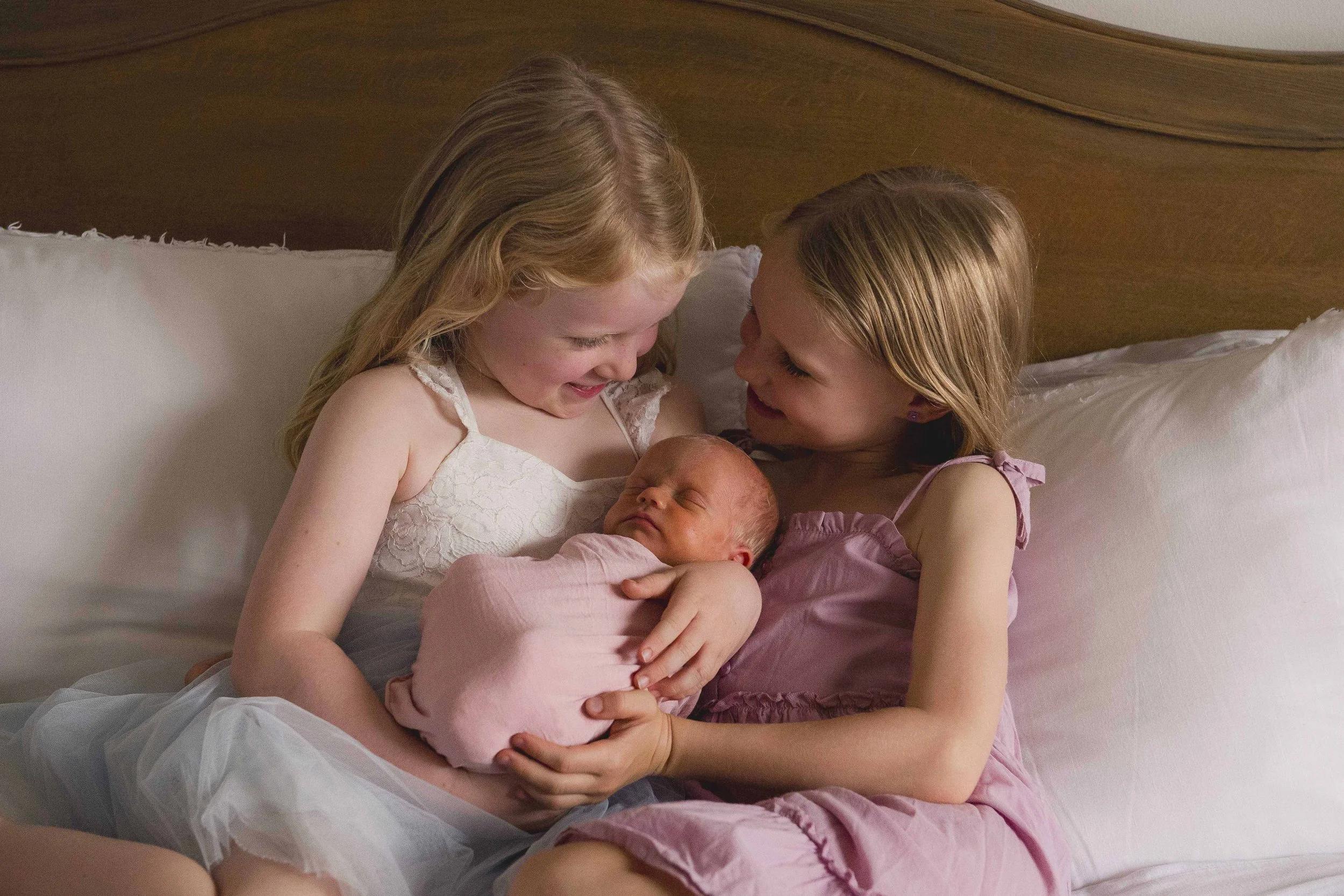 Two young girls, one with blonde hair and one with light brown hair, sitting on a bed and smiling at each other while holding a newborn baby dressed in pink. The scene is warm and intimate, with a wooden headboard in the background.