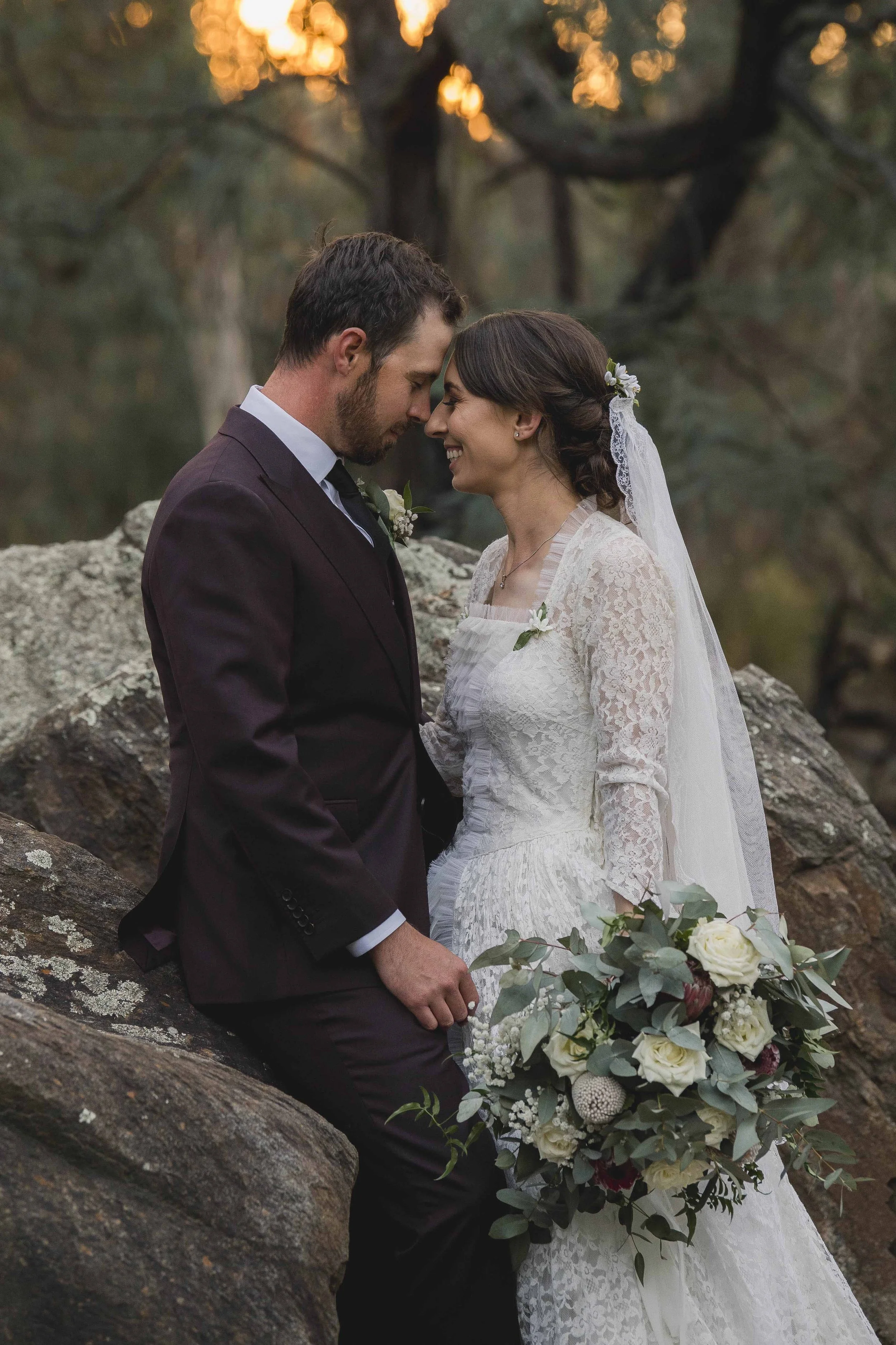 A bride and groom touching foreheads and smiling at each other outdoors, with the bride holding a bouquet of white roses and greenery, in a natural setting with rocks and trees in the background.