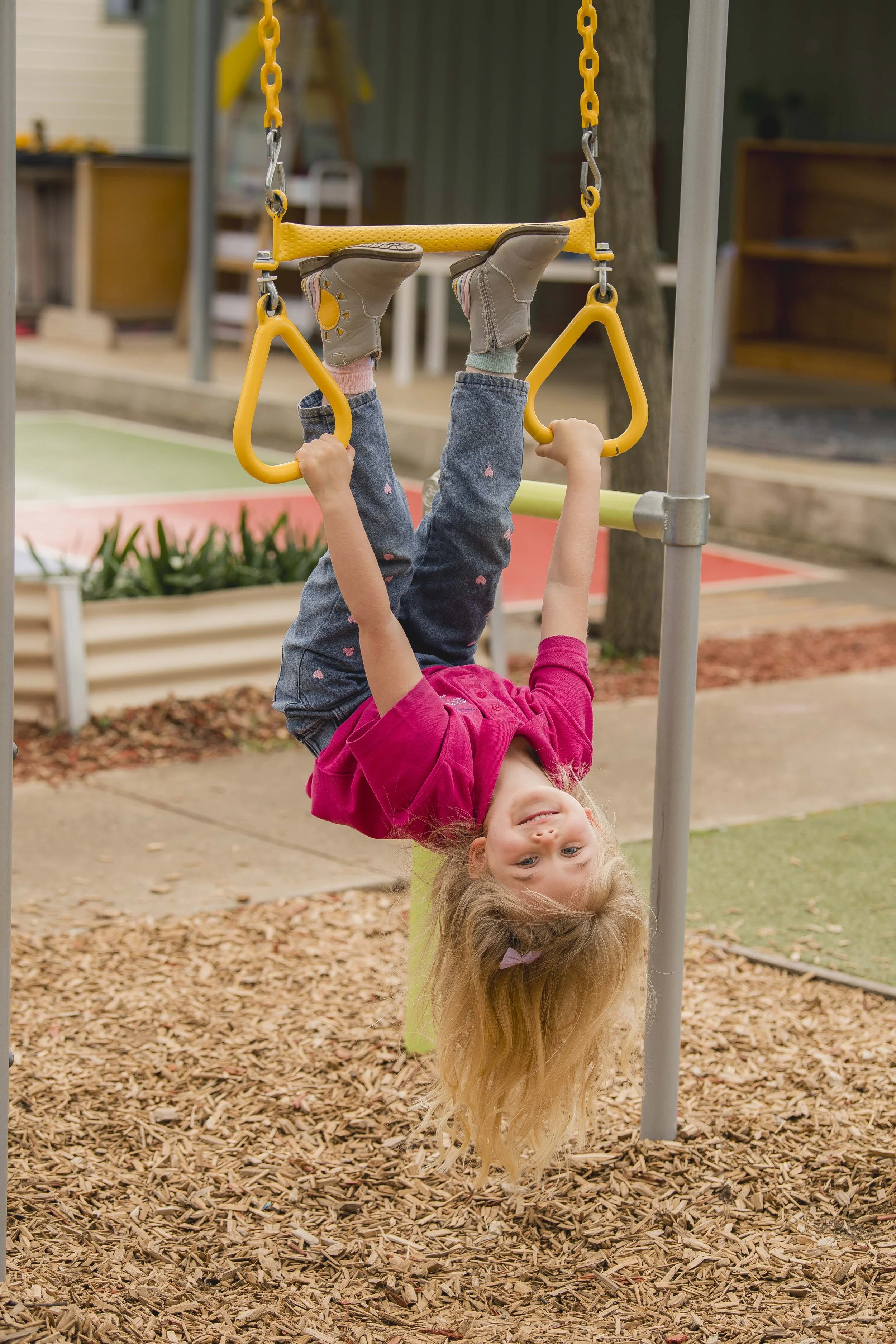 A young girl with long blonde hair hanging upside down from a playground swing, smiling and holding onto the swing's handles with her arms. She is wearing a pink shirt and jeans with pink heart patterns. The ground is covered with wood chips, and there are play structures and trees in the background.