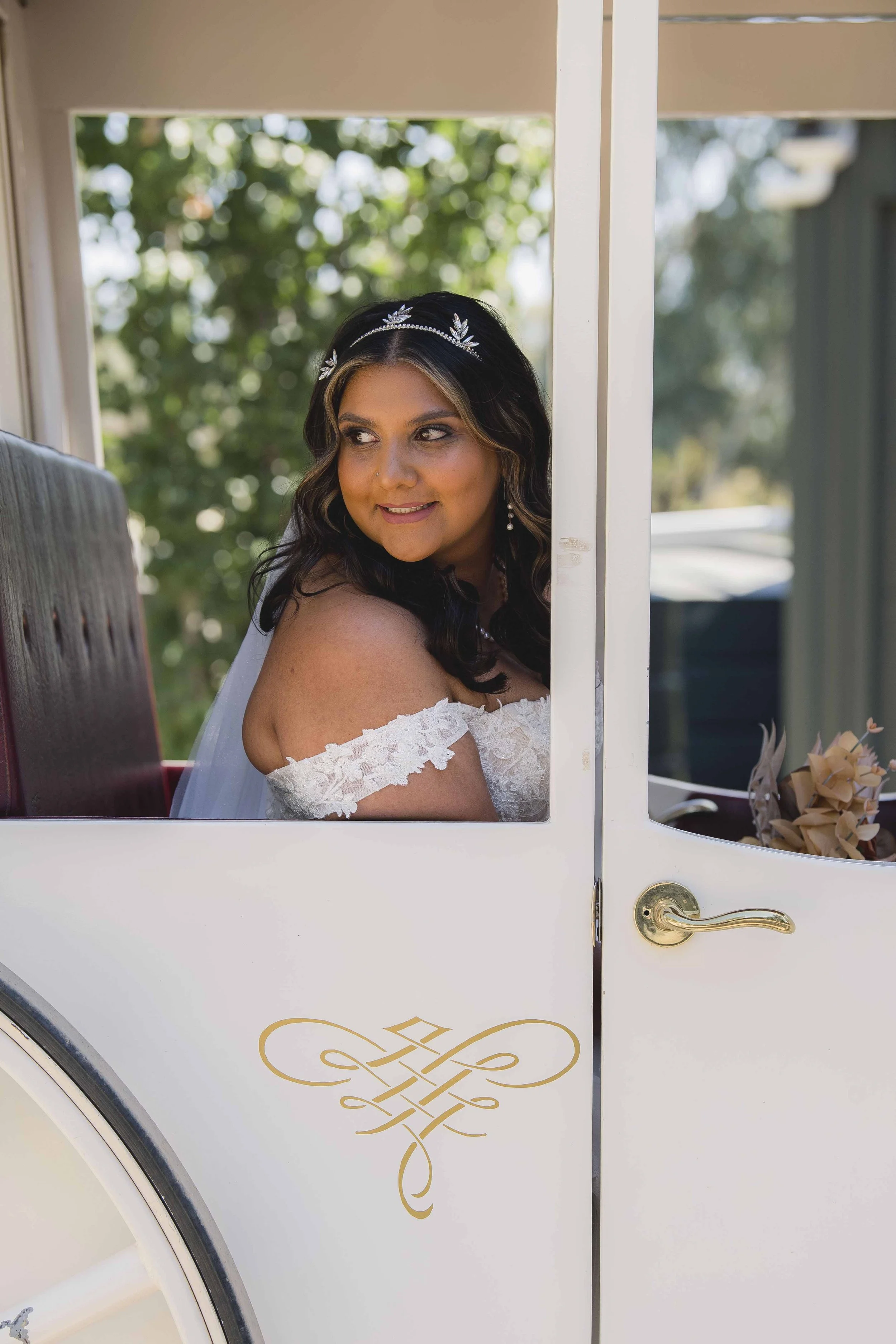 A woman in a wedding dress with lace sleeves and earrings, sitting inside a white carriage with gold decorations, with green trees in the background.