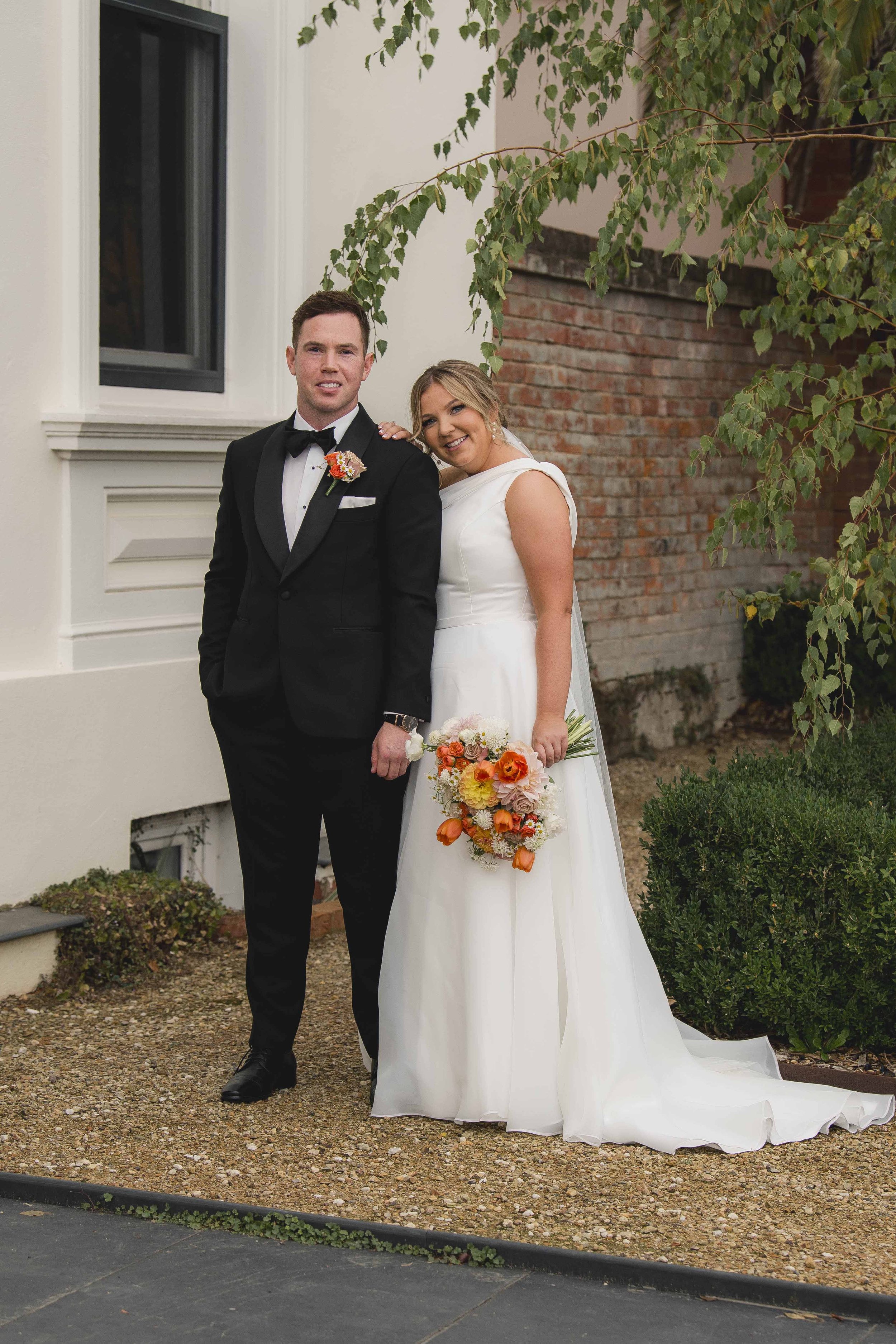A bride and groom standing outdoors, dressed in wedding attire, holding a bouquet of orange and white flowers. The bride has blonde hair styled in loose waves, and the groom has short dark hair, wearing a black tuxedo with a black bow tie and boutonniere, smiling beside a house with a brick wall and greenery.