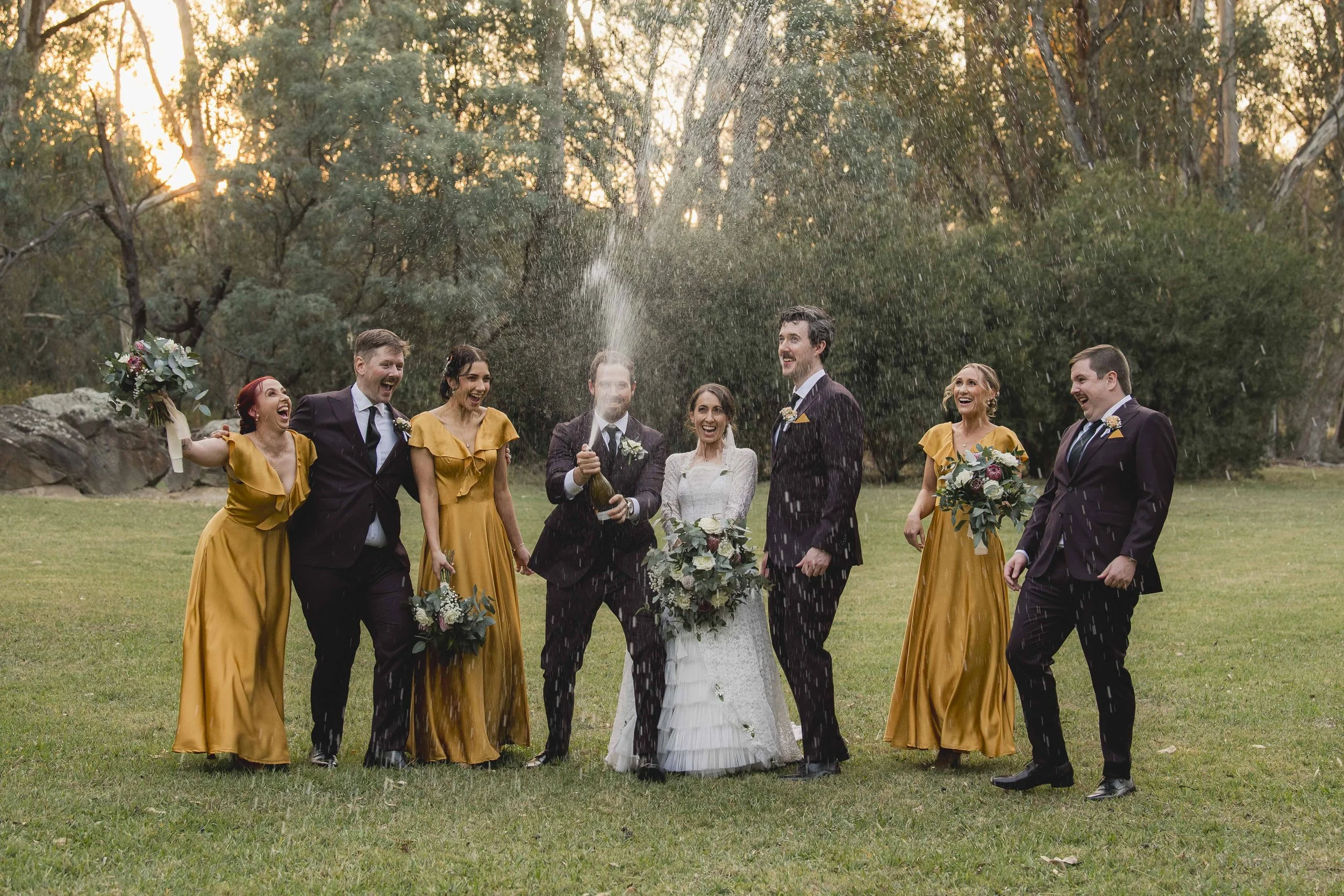 A group of  wedding guests, including the bride and groom, celebrating outdoors on grass with trees in the background during sunset. Four women in mustard yellow dresses and three men in dark suits are joyfully smiling, throwing and spraying champagn