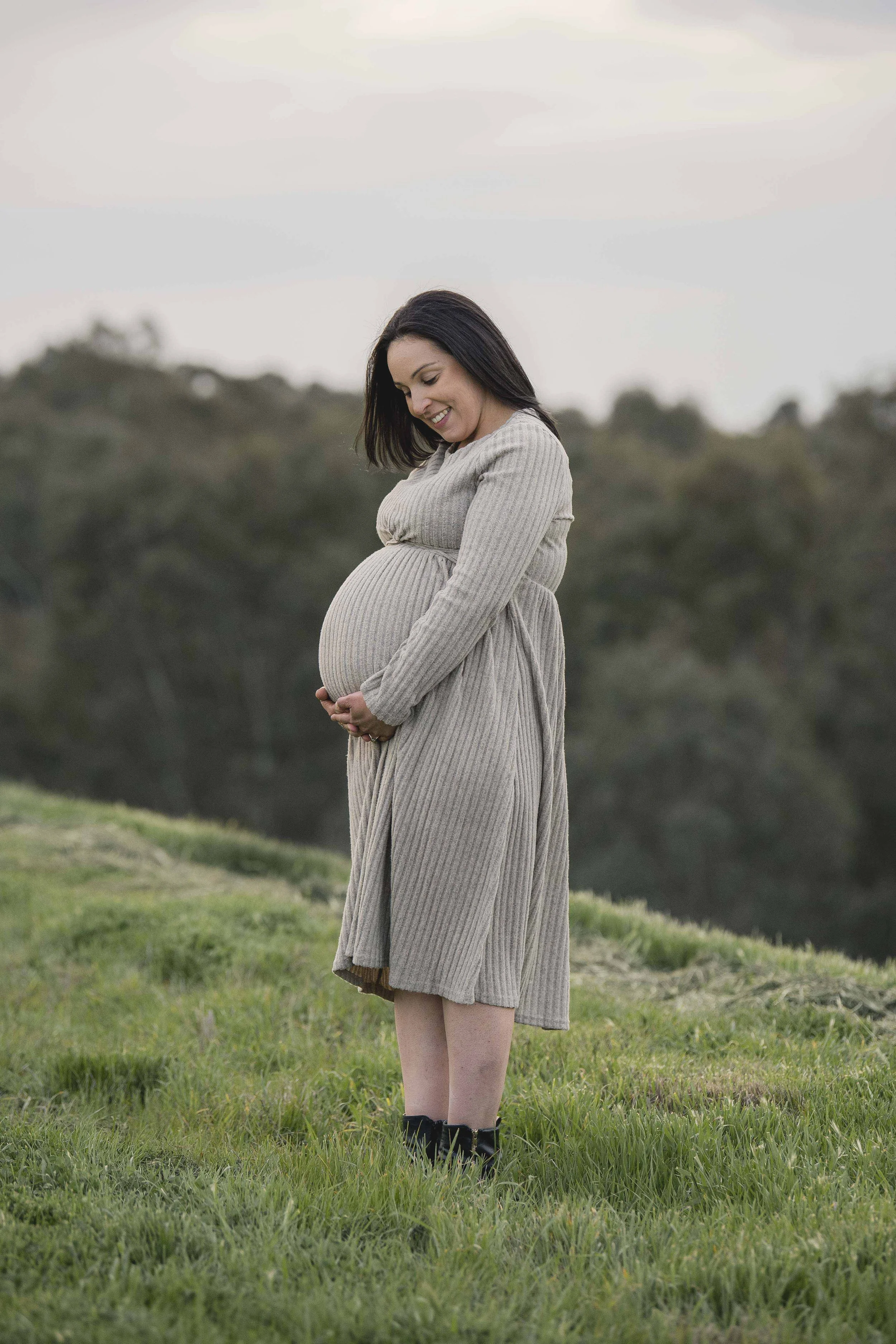 Pregnant woman standing outdoors in a grassy field, smiling, wearing a beige dress, black boots, and with dark hair, looking down at her belly.