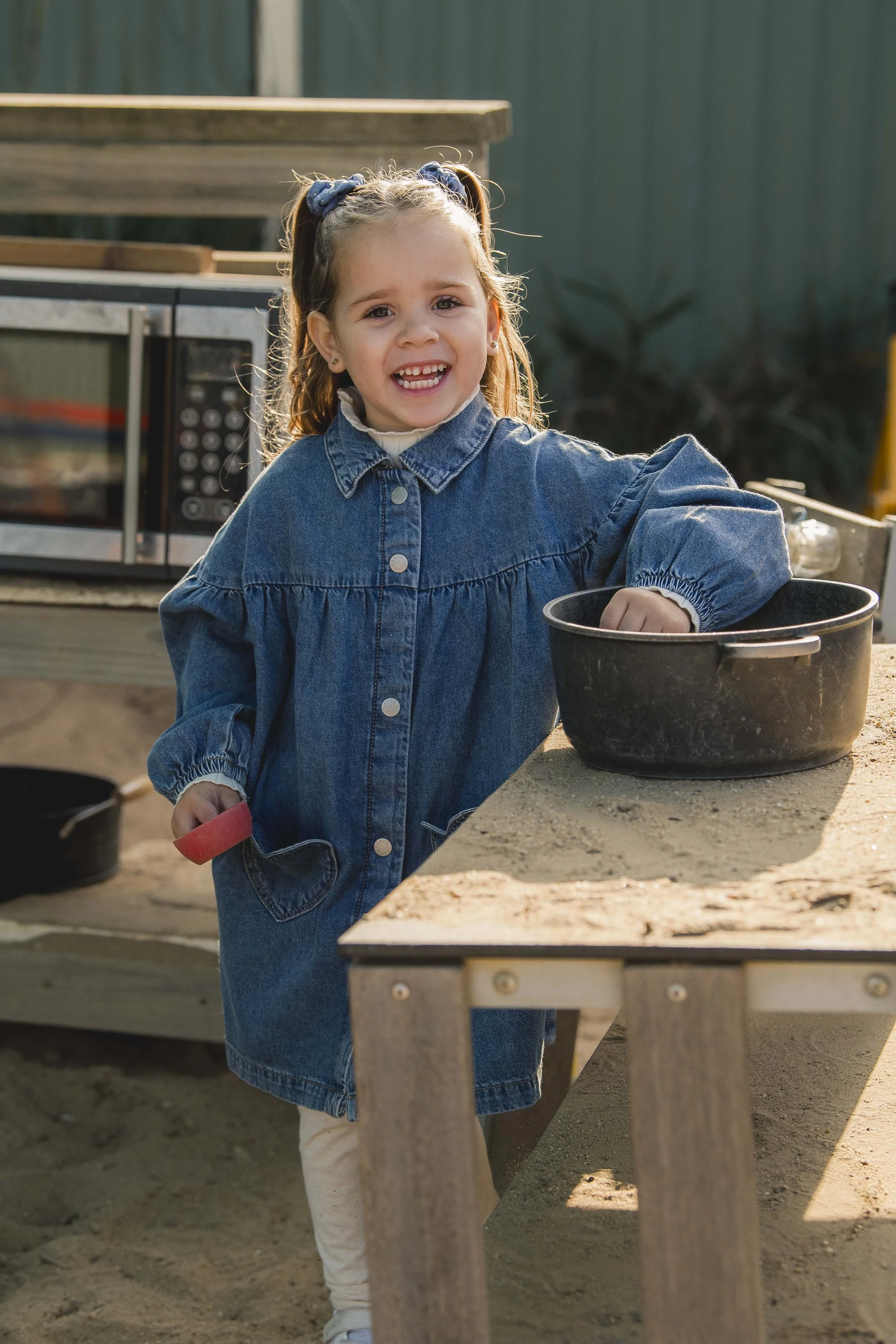 A young girl with a denim dress, smiling and standing outdoors, near a wooden table and a black pot, with a microwave oven in the background.