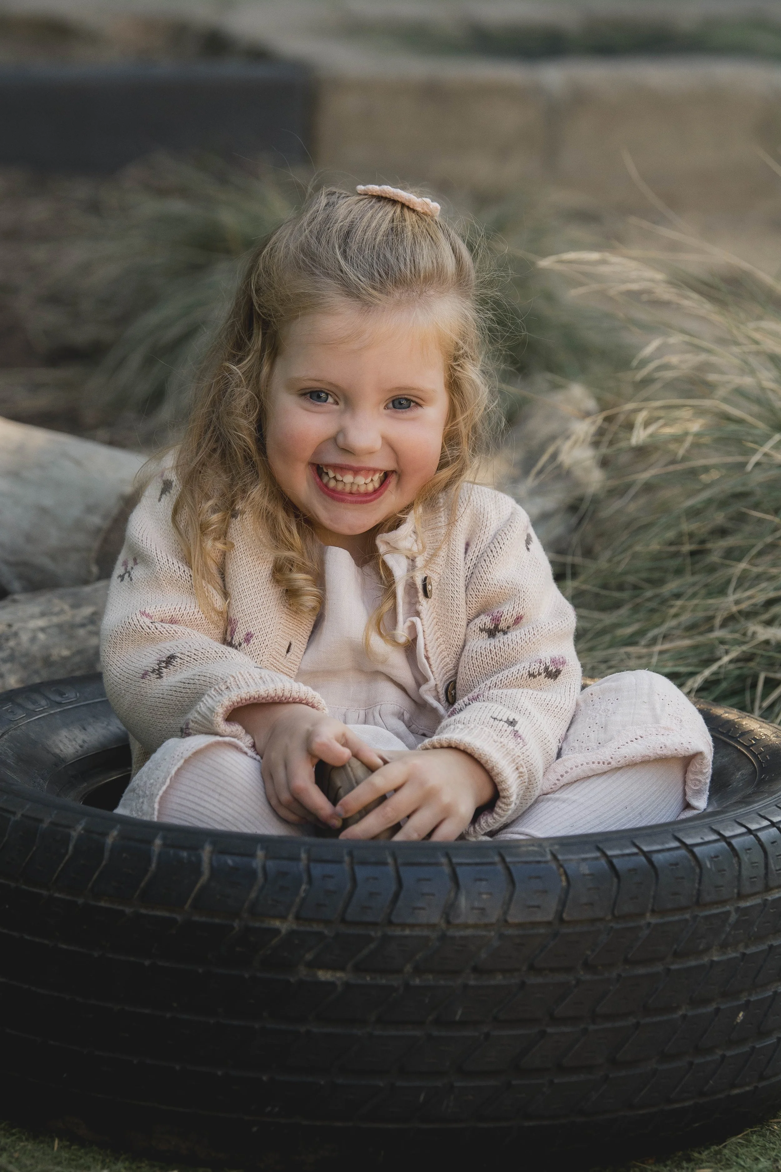 A young girl with curly blonde hair, wearing a beige sweater with small patterns, sitting inside a car tire which is on the ground outdoors. The girl is smiling and holding something in her hands.