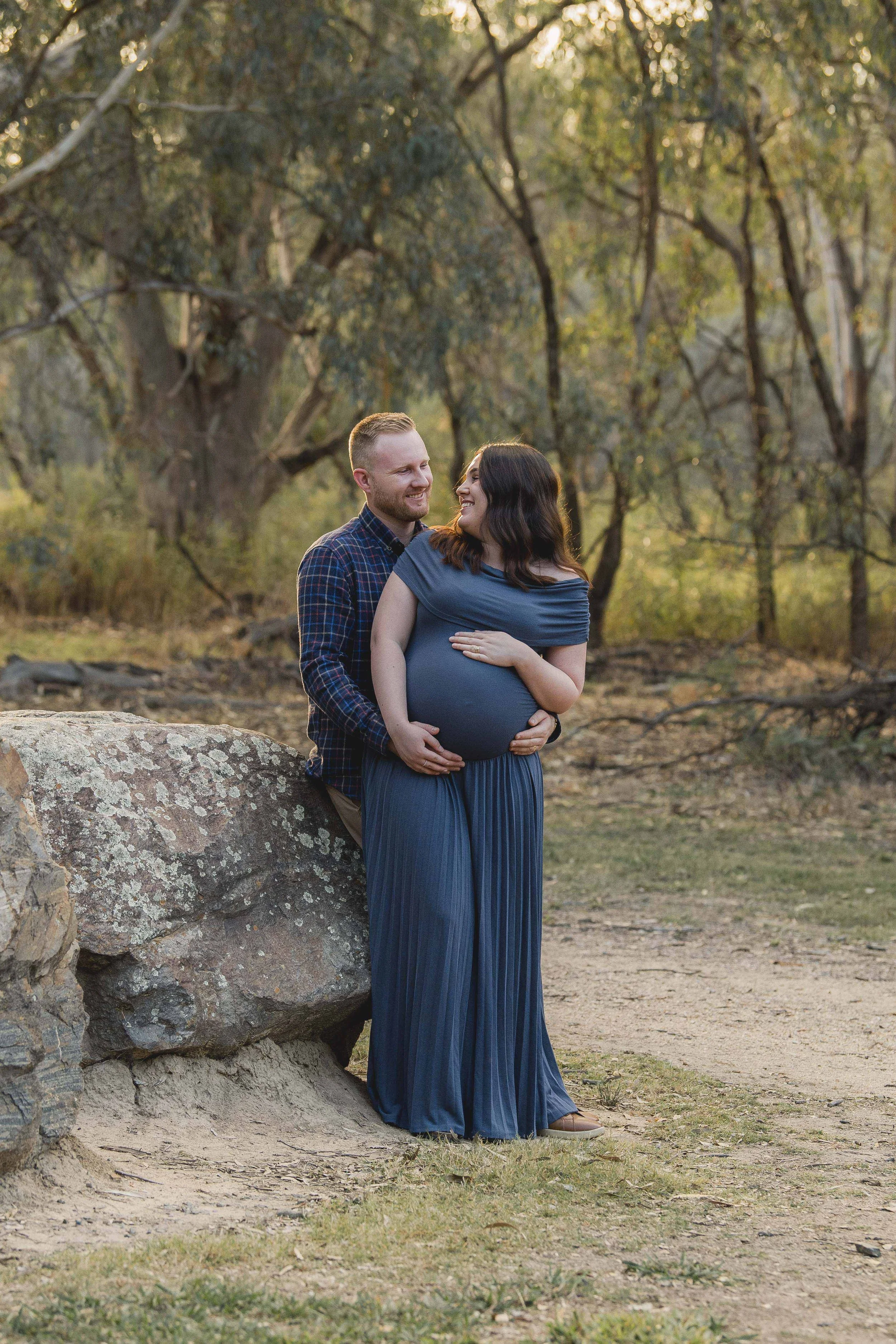 A couple standing outdoors near a large rock in a wooded area during sunset, with the woman visibly pregnant. The man has short blond hair and a beard, wearing a plaid shirt. The woman has long dark hair, wearing a blue dress that highlights her pregnancy, and they are smiling at each other.