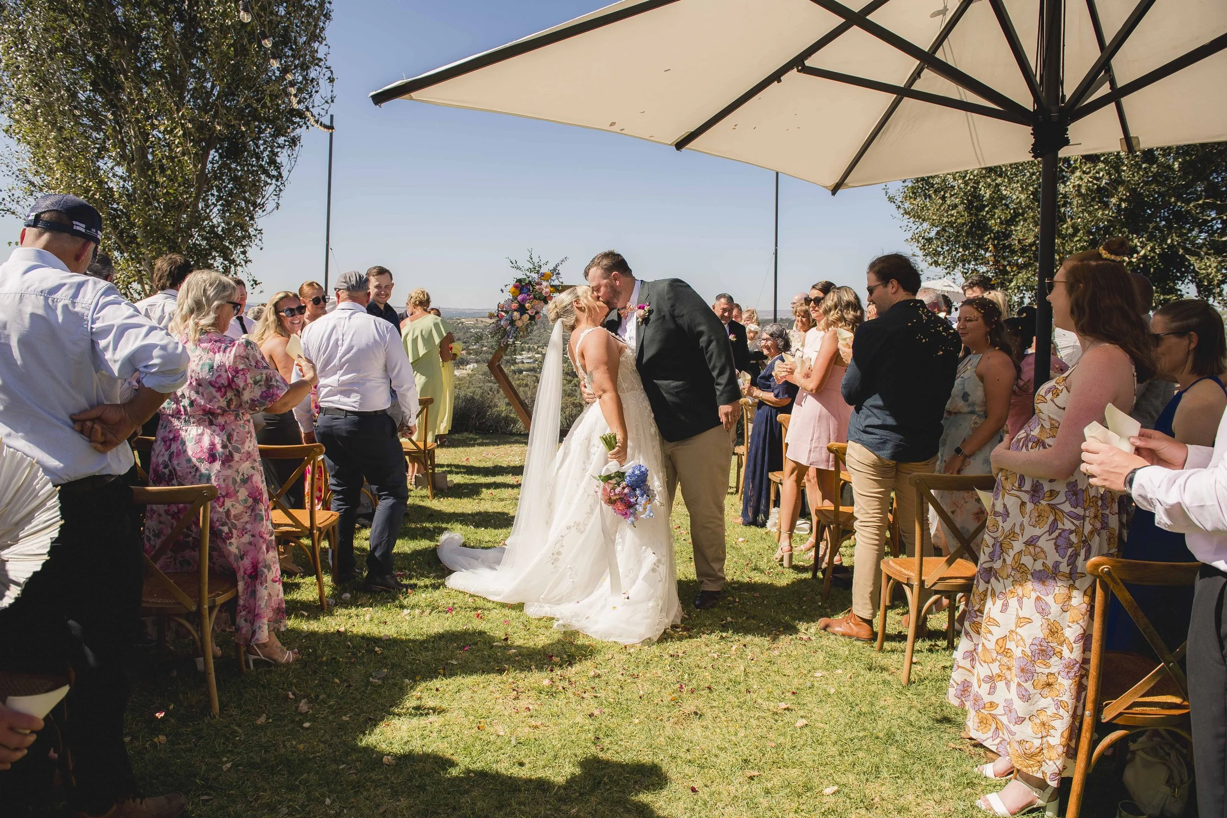 A bride and groom kissing at an outdoor wedding ceremony under a large umbrella, with guests seated on either side, on a sunny day with trees and a scenic view in the background.