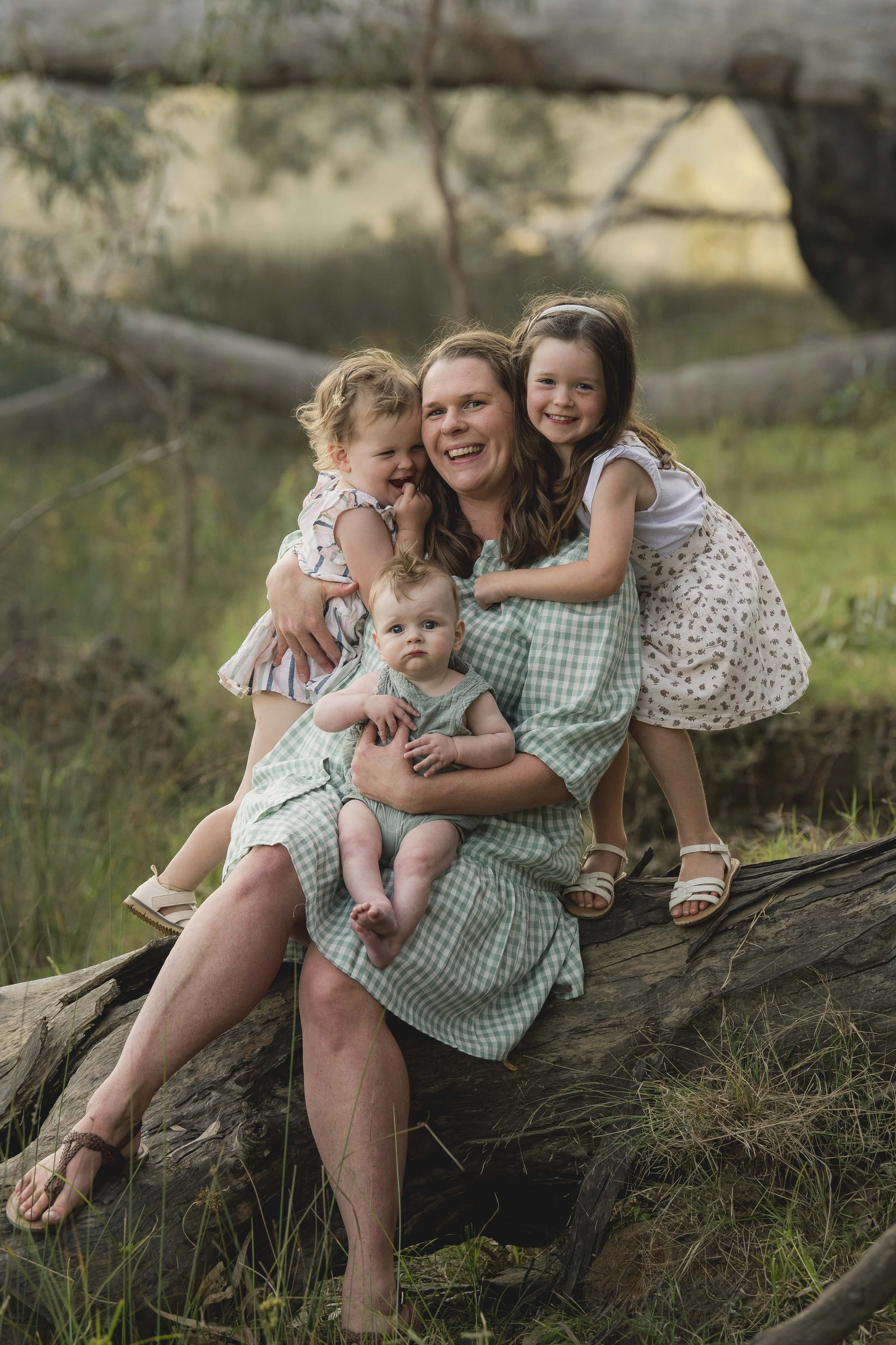 A woman and three young girls are sitting on a fallen tree in a grassy outdoor setting near a body of water with trees and a bridge in the background. They are smiling and appear to be enjoying nature.