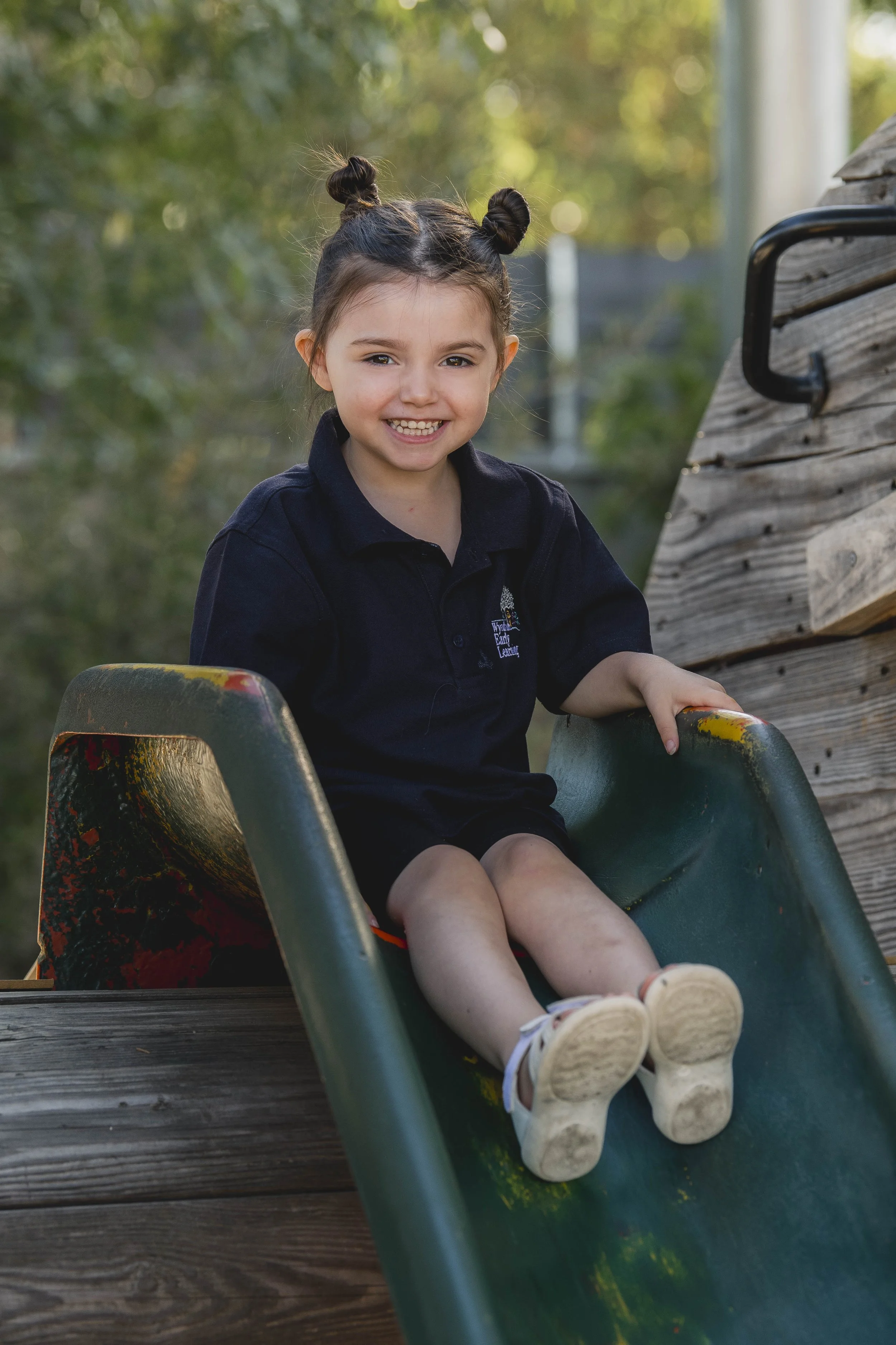 A young girl with brown hair styled into two space buns, wearing a navy blue polo shirt, smiling while sitting on a playground slide.