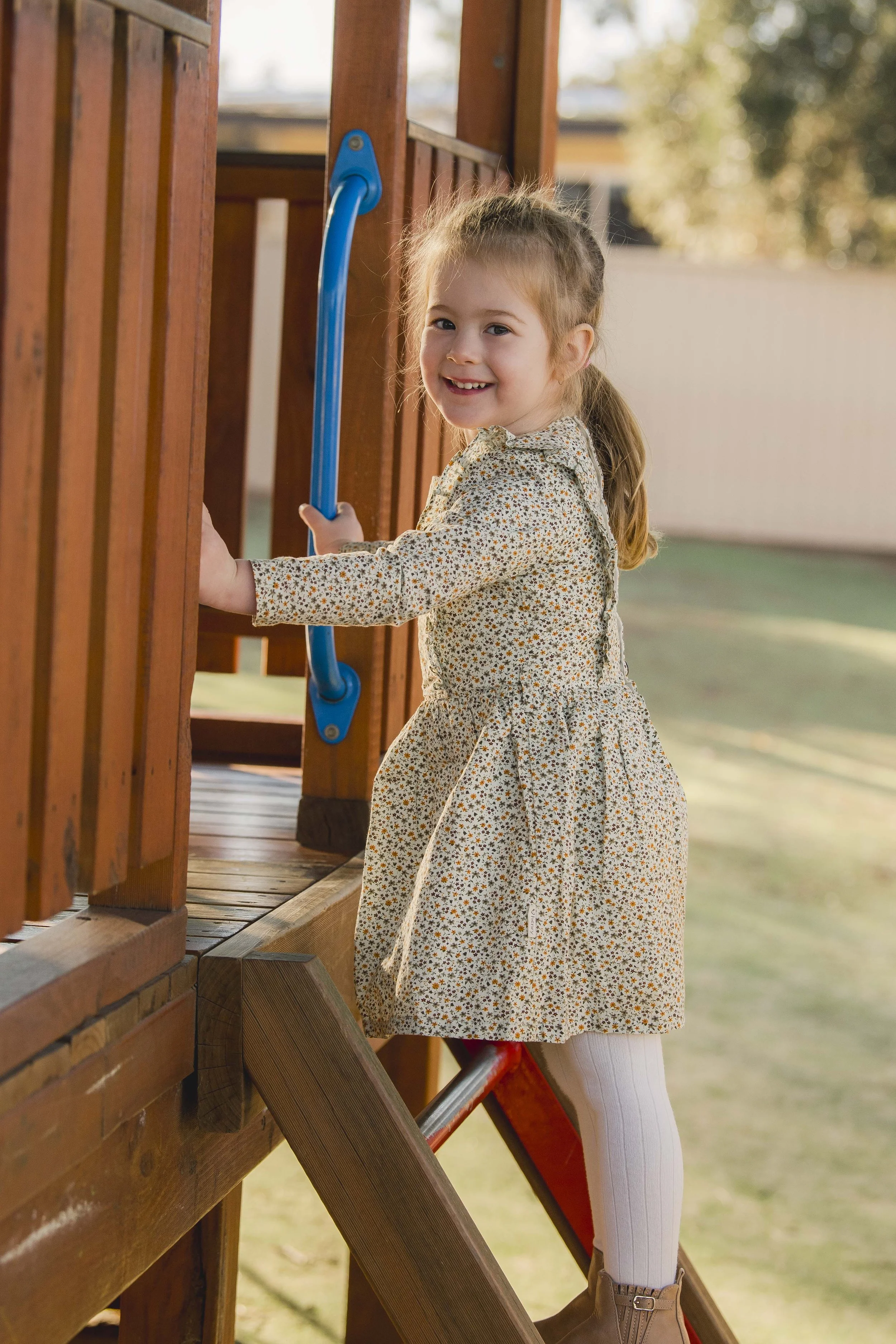 A young girl with light brown hair tied back, wearing a beige floral dress, white tights, and beige boots, smiling while climbing a wooden play structure outdoors. The background is blurred with trees and a sky.