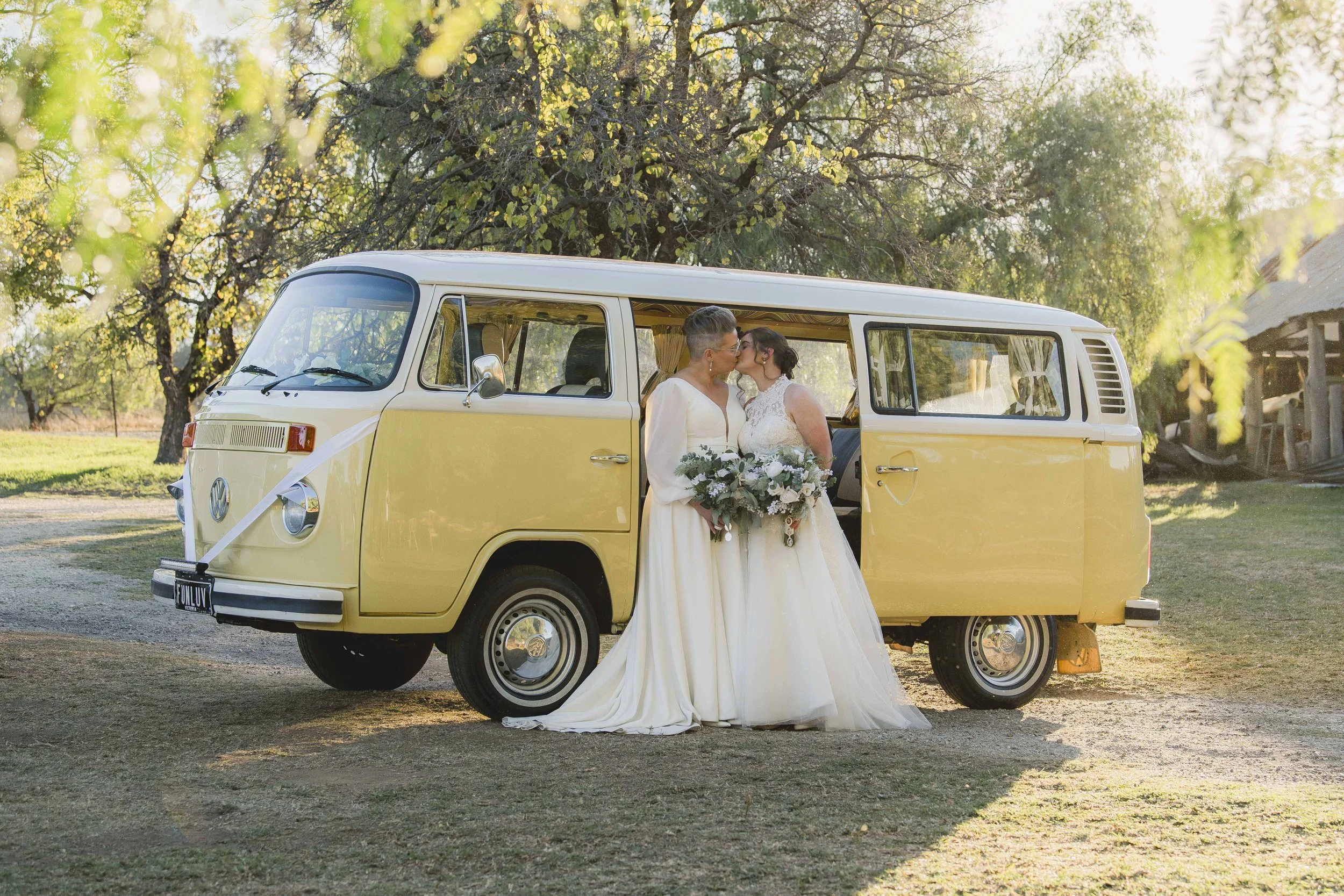 Two women in wedding dresses sharing a kiss inside a vintage yellow Volkswagen camper van parked outdoors among trees.